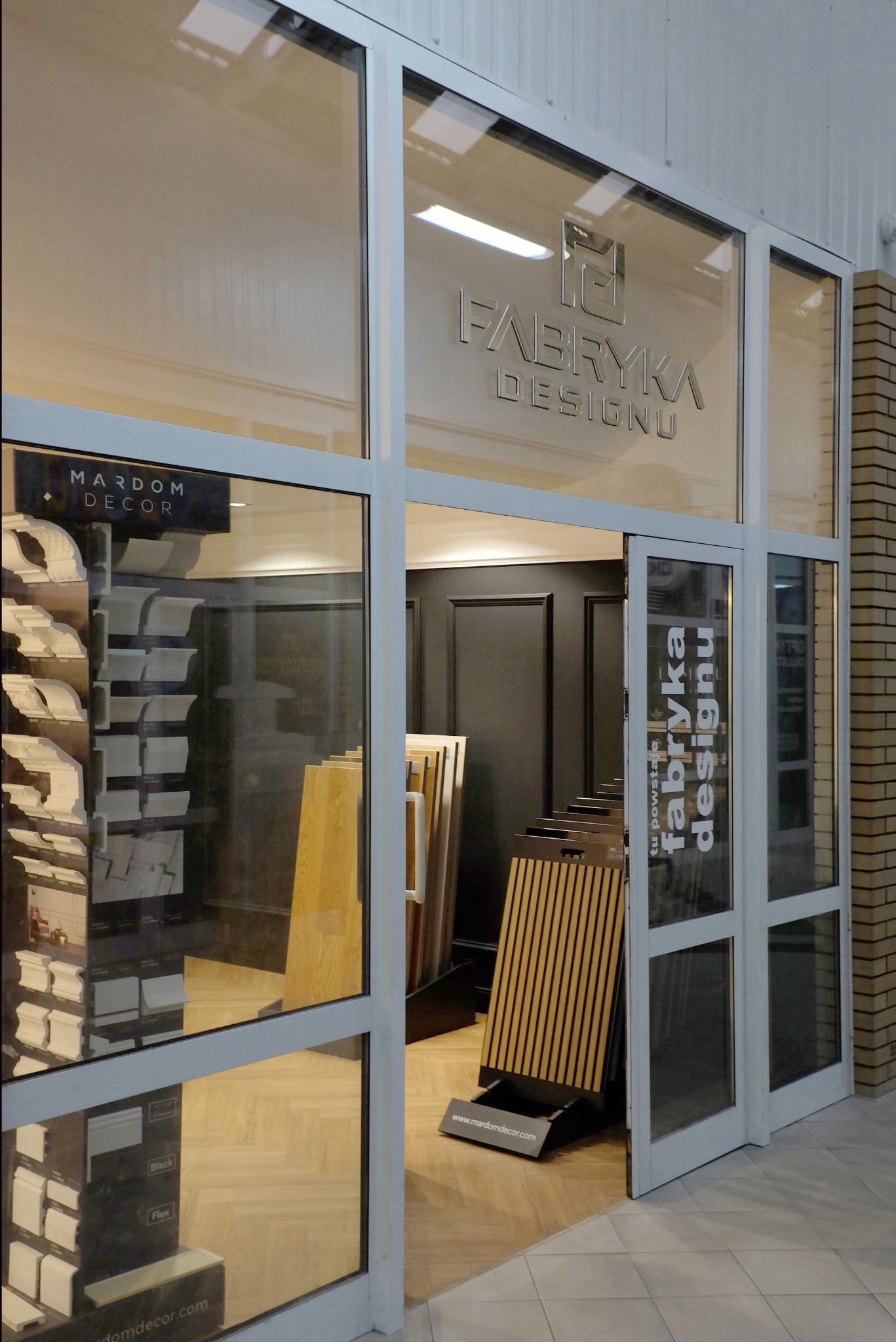Wejście do salonu Fabryka Designu z ekspozycją listew przypodłogowych Mardom Decor i paneli ściennych, widoczne przez szklane drzwi.