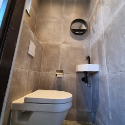 Piotr Szczepkowski usługi budowlane - Nowoczesna łazienka z szarymi płytkami, minimalistyczną umywalką z czarną armaturą, okrągłym lustrem i podwieszaną toaletą.