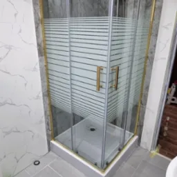 Narożna kabina prysznicowa z złotymi profilami i poziomymi pasami na szkle, zamontowana w łazience wykończonej płytkami imitującymi marmur i szarą terakotą.