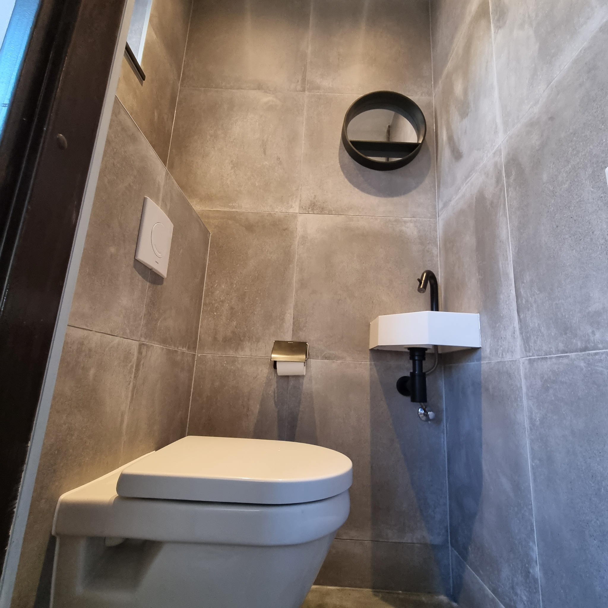 Nowoczesna łazienka z szarymi płytkami, minimalistyczną umywalką z czarną armaturą, okrągłym lustrem i podwieszaną toaletą.