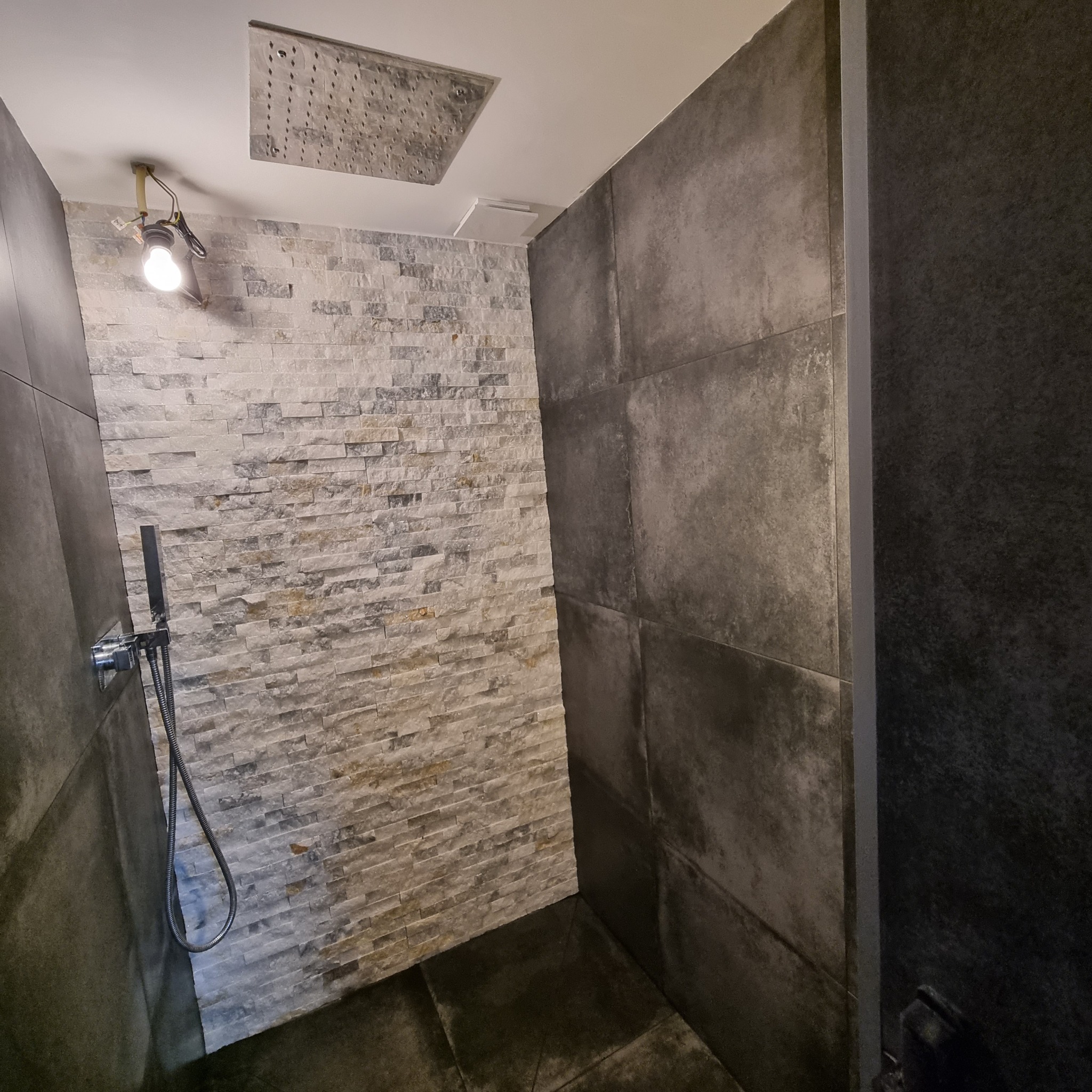 Nowoczesna kabina prysznicowa z szarą, strukturalną ścianą i białą ścianą z kamienia dekoracyjnego, deszczownica sufitowa, widoczna instalacja oświetleniowa.