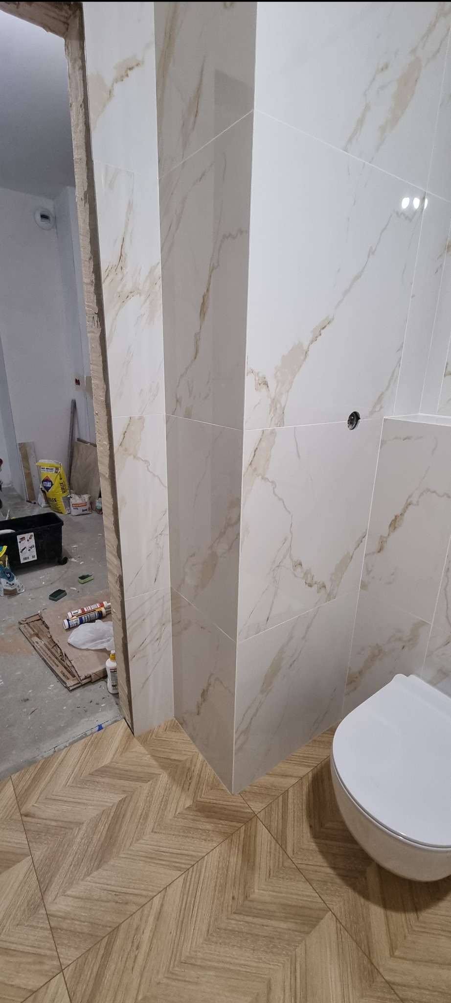 Białe płytki imitujące marmur z beżowymi żyłami na ścianie łazienki, układane w pionie, kontrastują z drewnopodobną podłogą ułożoną w jodełkę; widoczny fragment toalety i materiały budowlane w tle,...