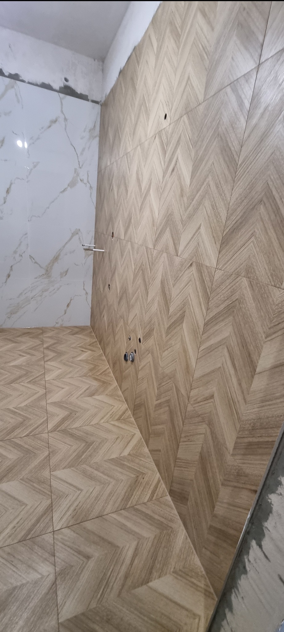 Ukończona ściana i podłoga w łazience z drewnopodobnych płytek ułożonych we wzór jodełki, widoczne otwory na instalację hydrauliczną i fragment ściany z białych marmurowych płytek.