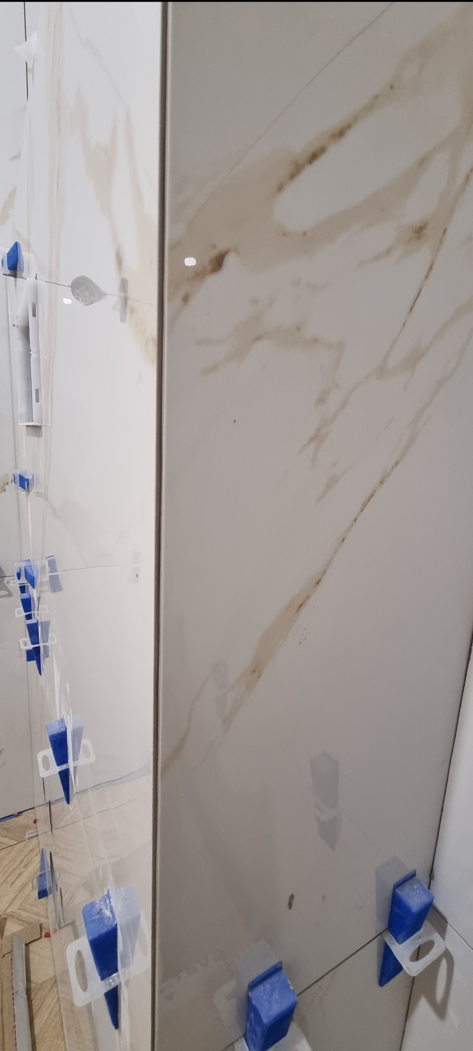 Układanie płytek imitujących marmur na ścianie, z użyciem niebieskich klinów poziomujących, widoczne linie poziomowania