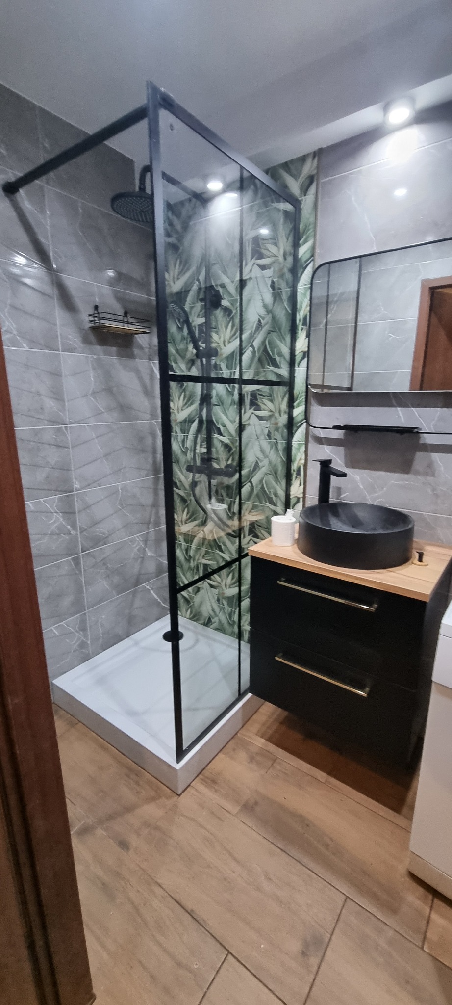 Nowoczesna łazienka z kabiną prysznicową typu walk-in z czarnymi profilami, umywalką nablatową w kolorze czarnym na szafce z blatem drewnopodobnym i tapetą z motywem liści.