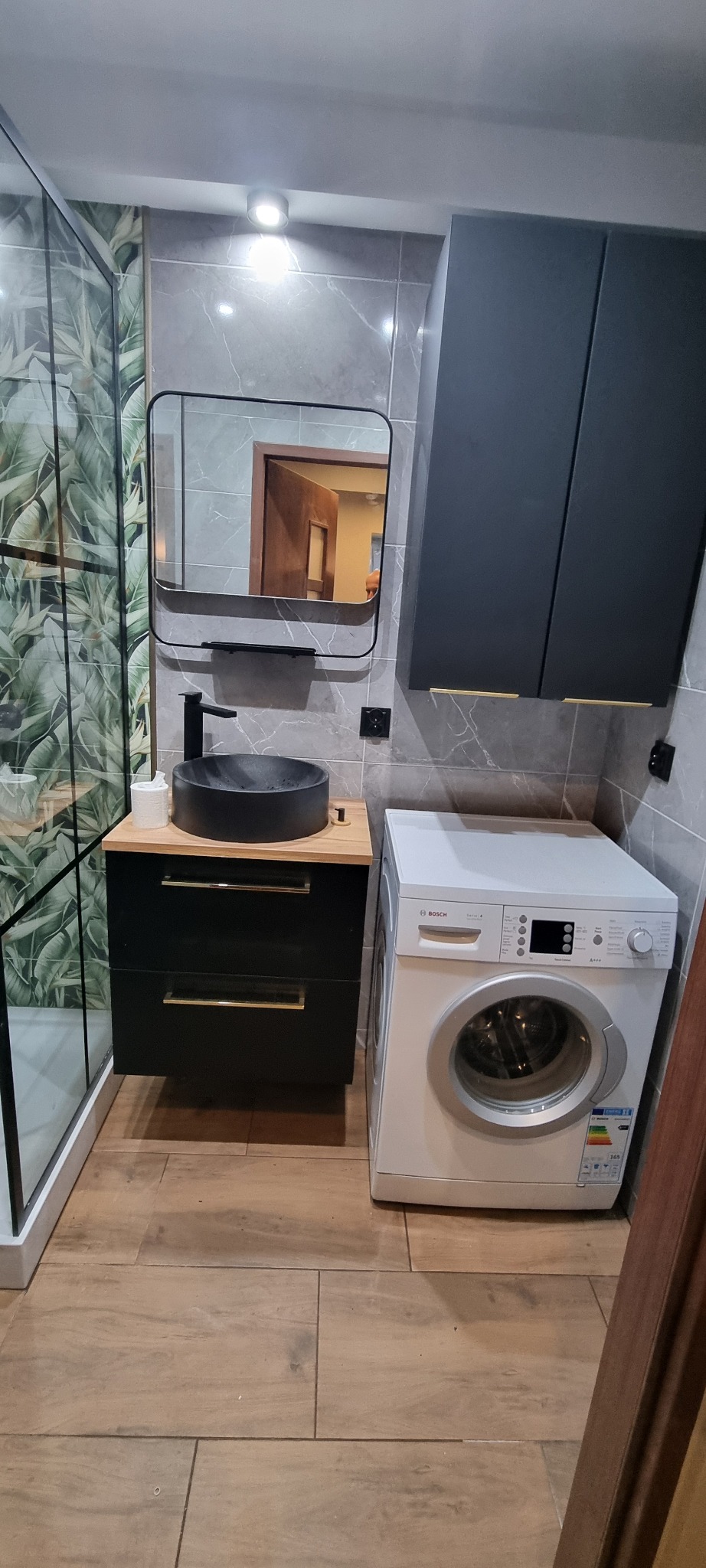 Nowoczesna łazienka z kabiną prysznicową w tropikalne liście, czarną umywalką nablatową, szafką z drewnianym blatem i złotymi uchwytami oraz pralką Bosch Serie 4.