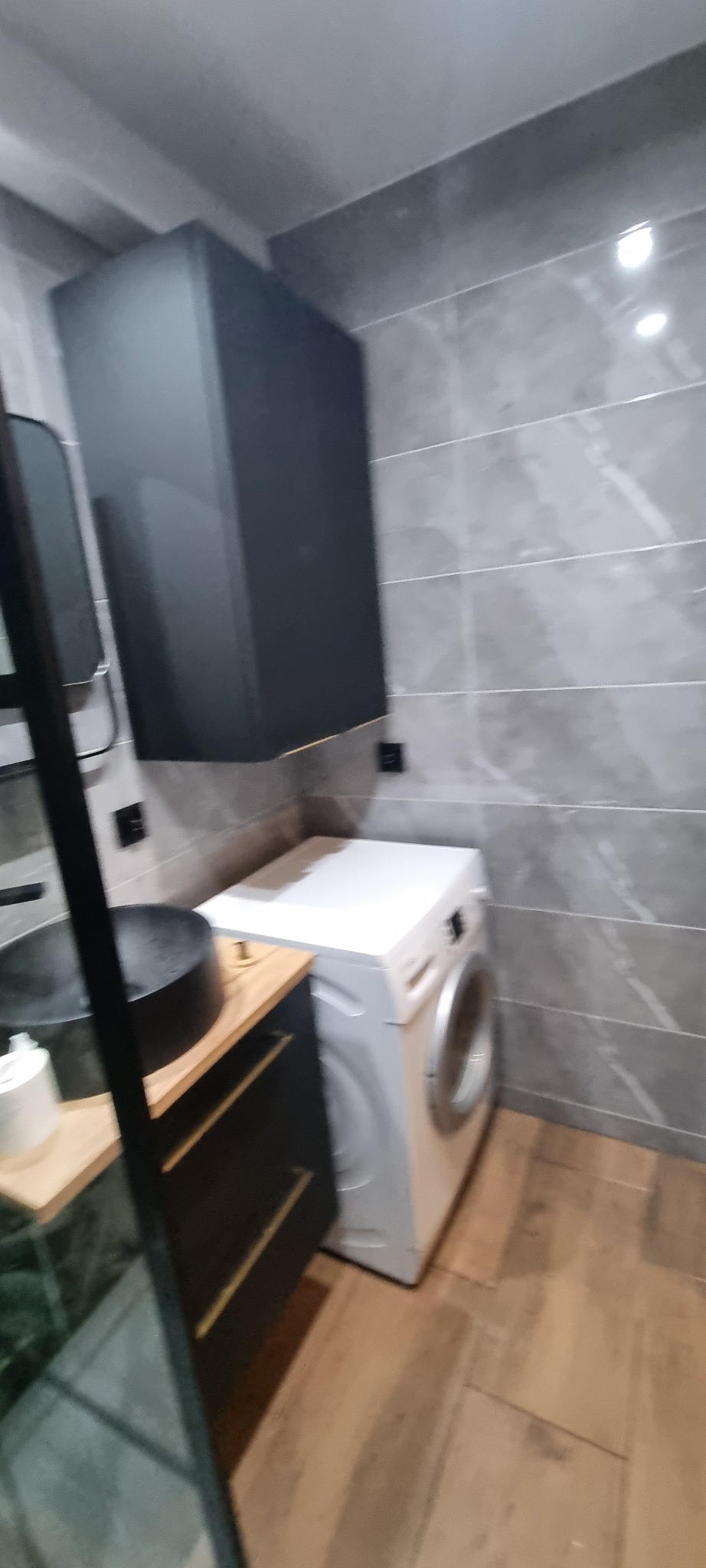 Nowoczesna łazienka z szarą płytką imitującą kamień, czarną szafką pod umywalką z drewnianym blatem, czarną wiszącą szafką i białą pralką.