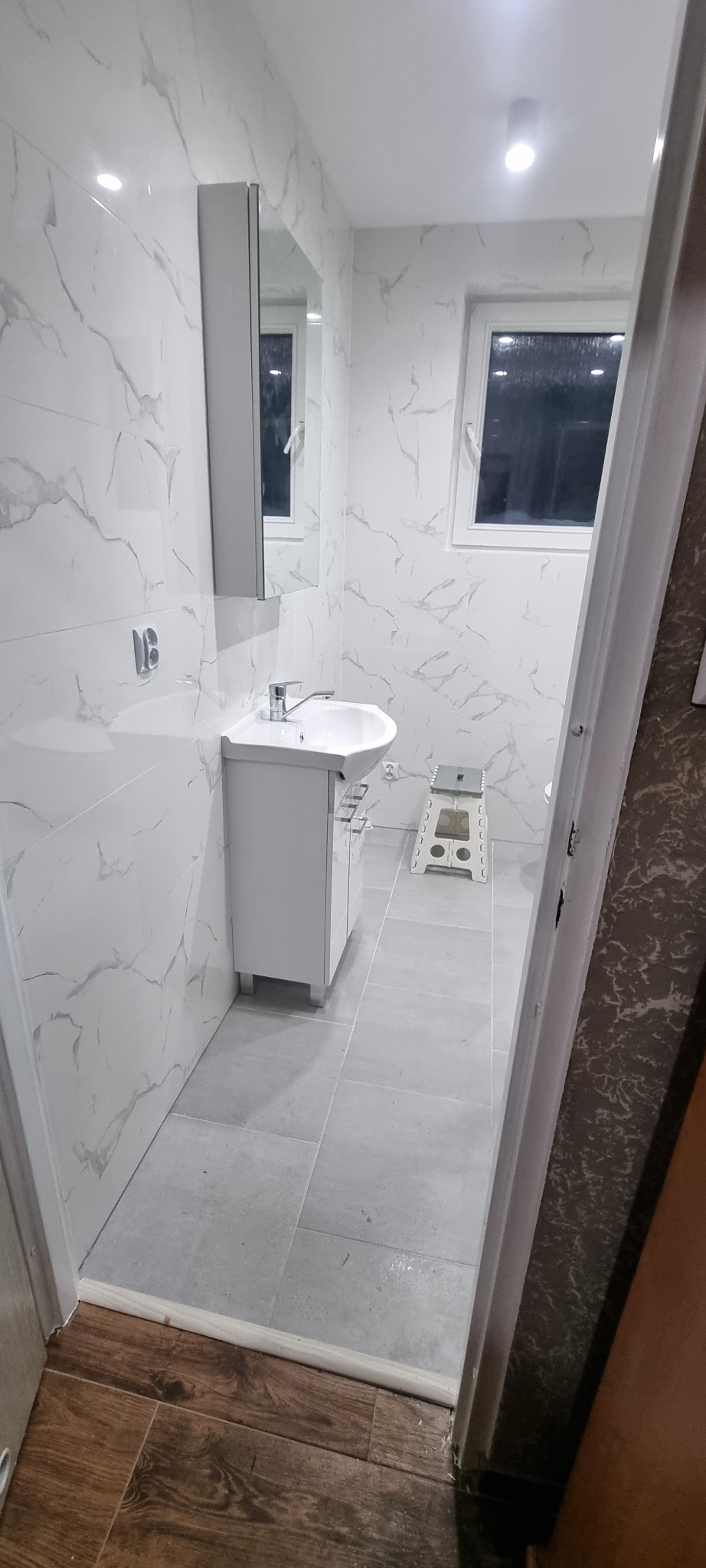 Nowoczesna łazienka w bieli i szarości z marmurkowymi płytkami, umywalką z szafką, lustrem w szafce i stołkiem drabinkowym w tle, widok zza otwartych drzwi.