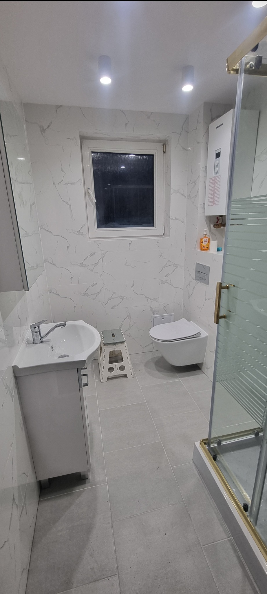 Nowoczesna łazienka wykończona w bieli i szarości, z marmurową teksturą na ścianach, wiszącą toaletą, umywalką z szafką, kabiną prysznicową ze złotymi detalami i drabinką-stołkiem.