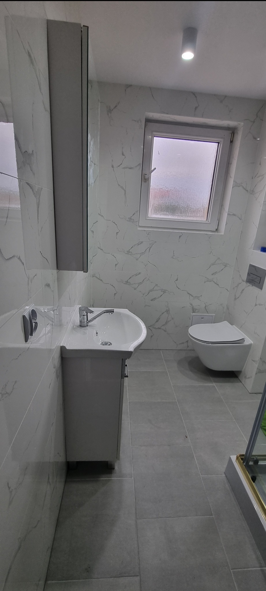 Nowoczesna łazienka w odcieniach szarości i bieli z umywalką na szafce, podwieszaną toaletą i marmurowymi płytkami na ścianach.