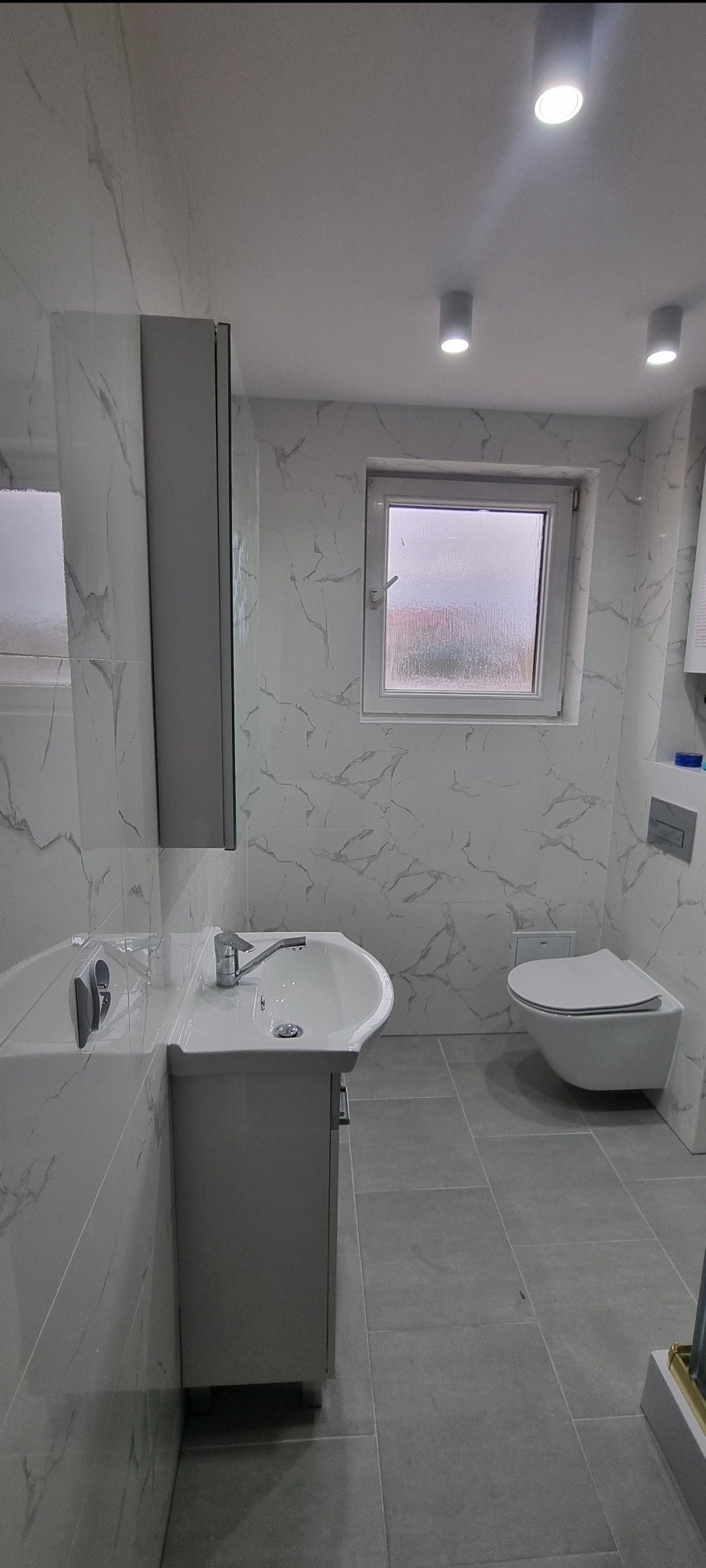 Nowoczesna łazienka z umywalką na szafce, wiszącą toaletą i marmurowymi płytkami na ścianach, oświetlona punktowymi lampami sufitowymi.