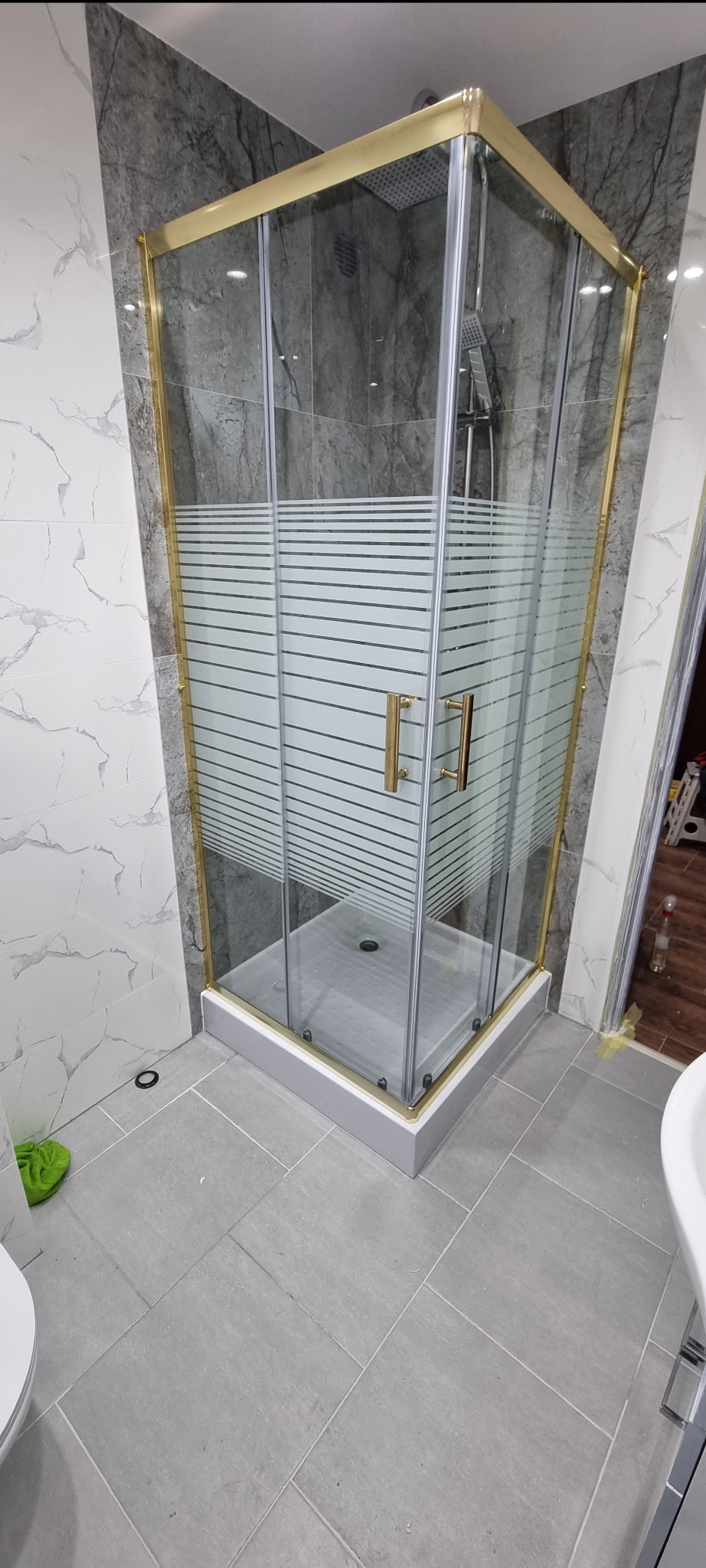 Narożna kabina prysznicowa z złotymi profilami i poziomymi pasami na szkle, zamontowana w łazience wykończonej płytkami imitującymi marmur i szarą terakotą.