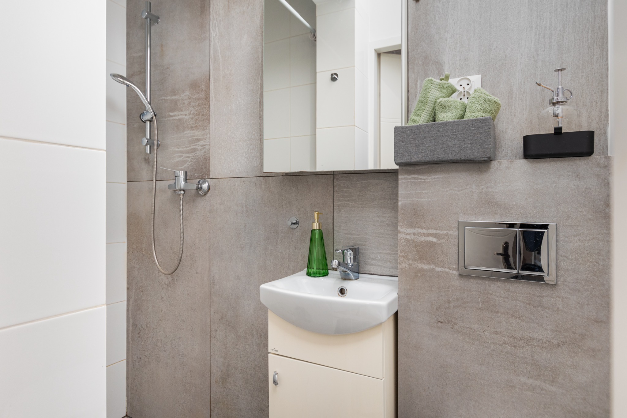 Jasna łazienka z kabiną prysznicową, umywalką z szafką, zielonym dozownikiem mydła, szarymi ręcznikami i armaturą w odcieniach srebra, na tle płytek imitujących kamień.