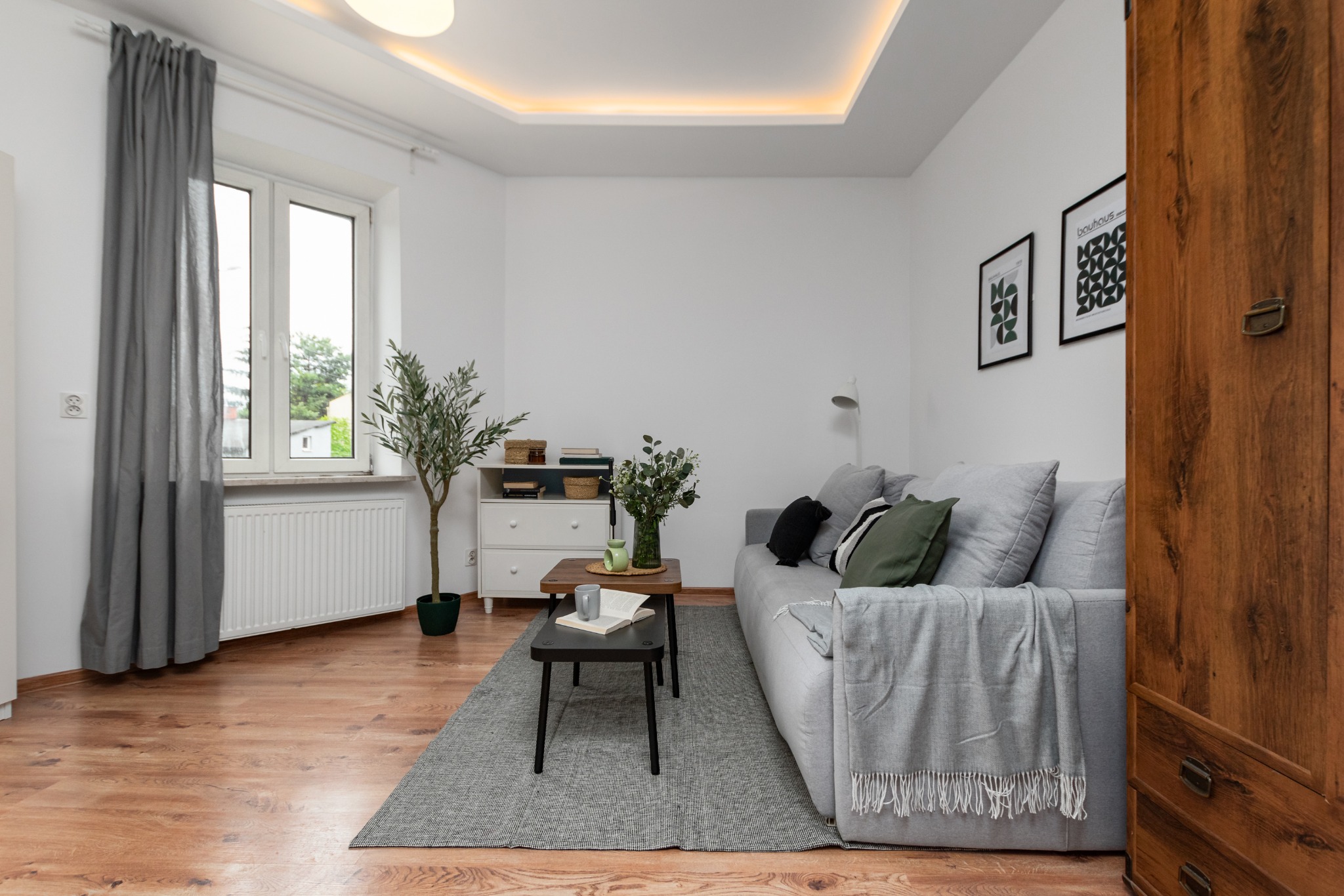 Jasne wnętrze salonu w Warszawie, z szarą sofą z poduszkami i pledem, dwoma stolikami kawowymi, komodą z rośliną w doniczce i dekoracjami ściennymi, widziane z perspektywy wejścia.