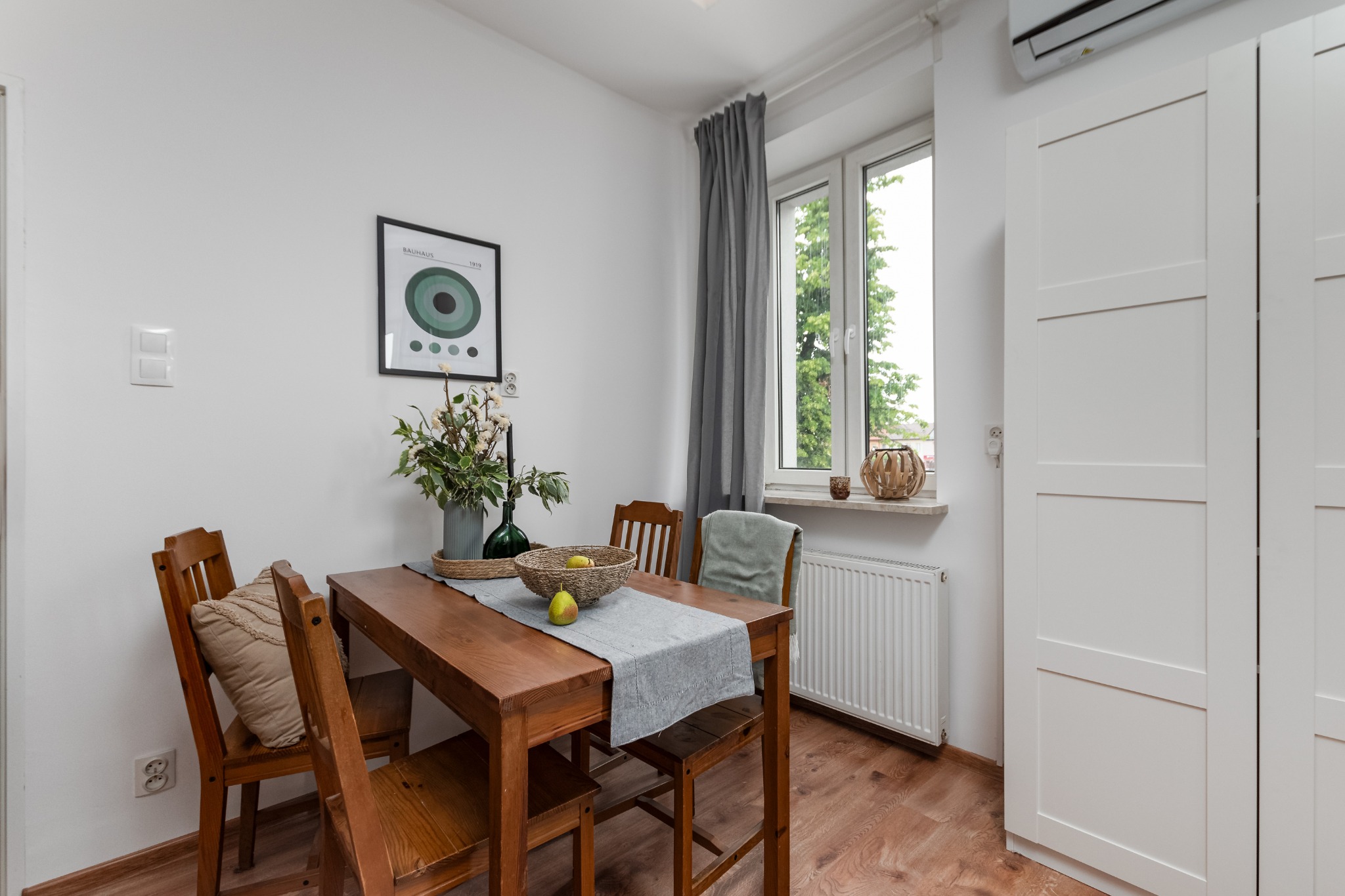 Jasna jadalnia z drewnianym stołem, na którym leży szary bieżnik, koszyk z owocami i wazon z kwiatami. W tle okno z szarą zasłoną, biały grzejnik i biała szafa.