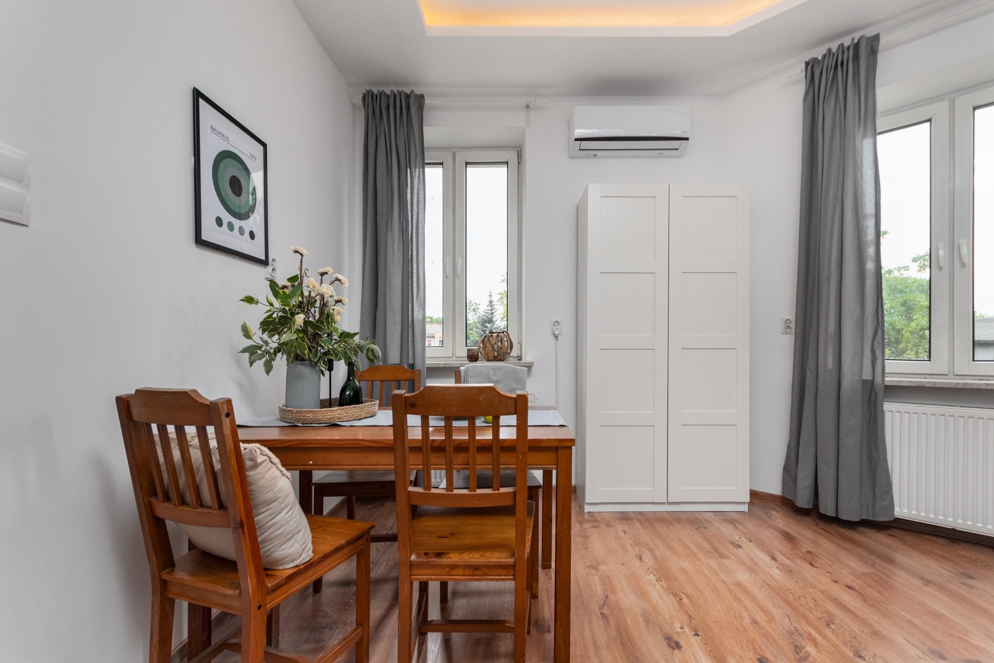 Jasna jadalnia z drewnianym stołem, krzesłami z poduszką, białymi szafkami, szarymi zasłonami i plakatem Bauhaus na ścianie.