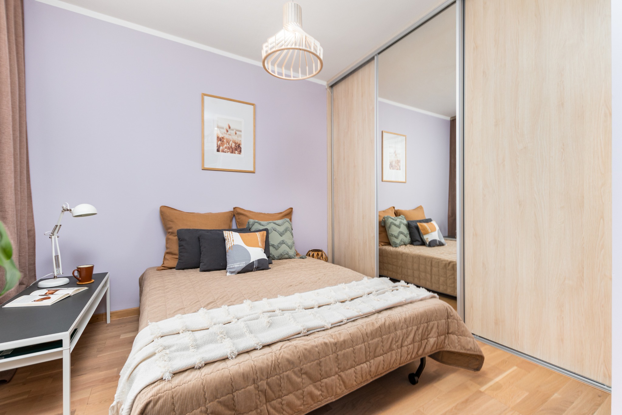 Jasna sypialnia z fioletowymi ścianami, łóżkiem z beżową narzutą i dekoracyjnymi poduszkami, drewnianą podłogą oraz szafą z lustrzanymi drzwiami przesuwnymi.