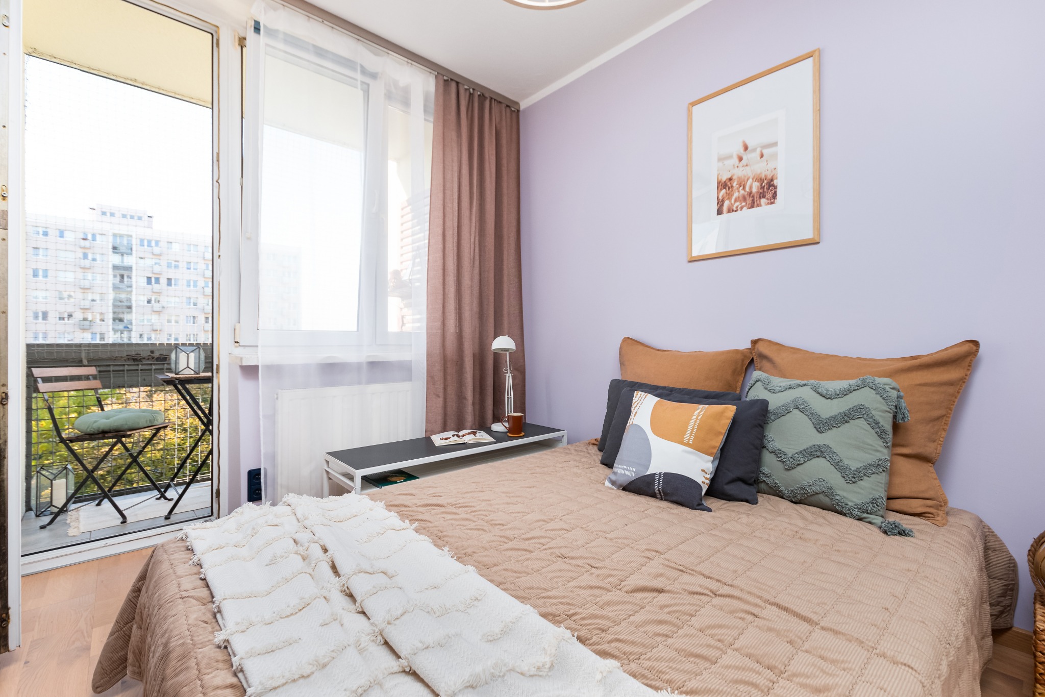 Jasna sypialnia z łóżkiem, stolikiem nocnym i balkonem z widokiem na miasto.