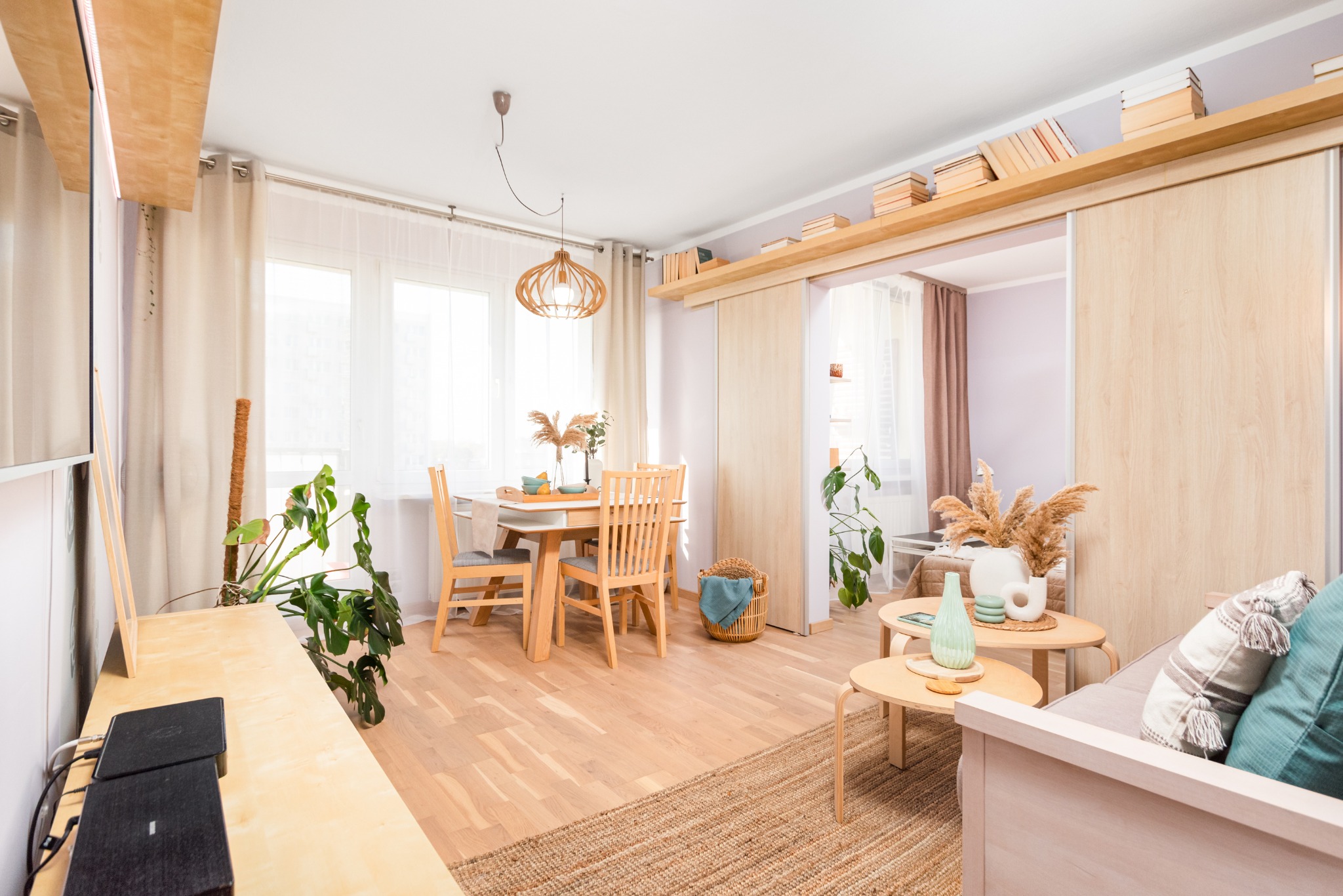 Jasne wnętrze mieszkania z drewnianym stołem, krzesłami i dekoracjami z suszonych traw, oświetlone naturalnym światłem wpadającym przez okno.