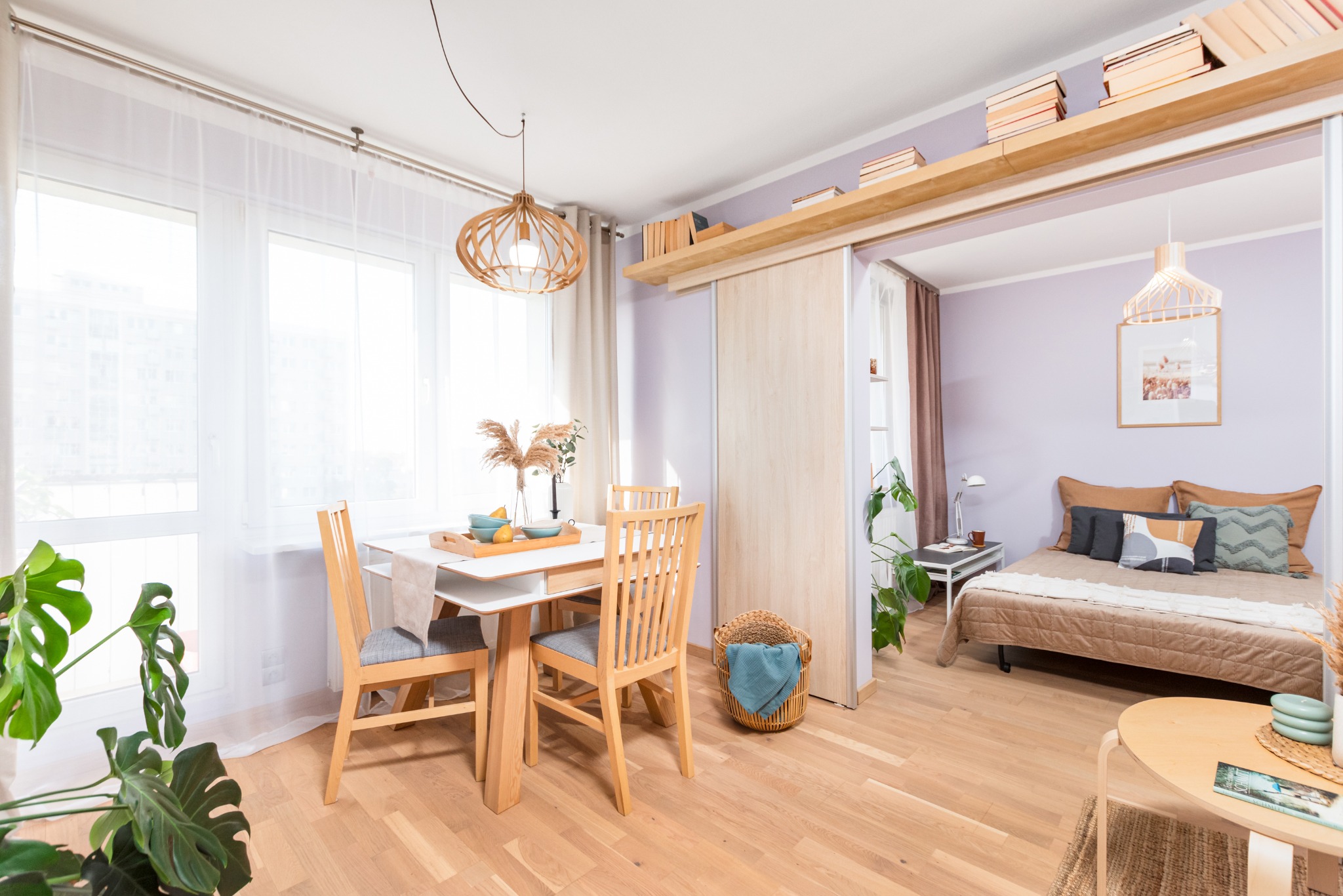 Jasne, przestronne wnętrze mieszkania z otwartą przestrzenią jadalni i sypialni, drewniany stół z nakryciem i dekoracjami, widok na sypialnię z łóżkiem i półkami z książkami.