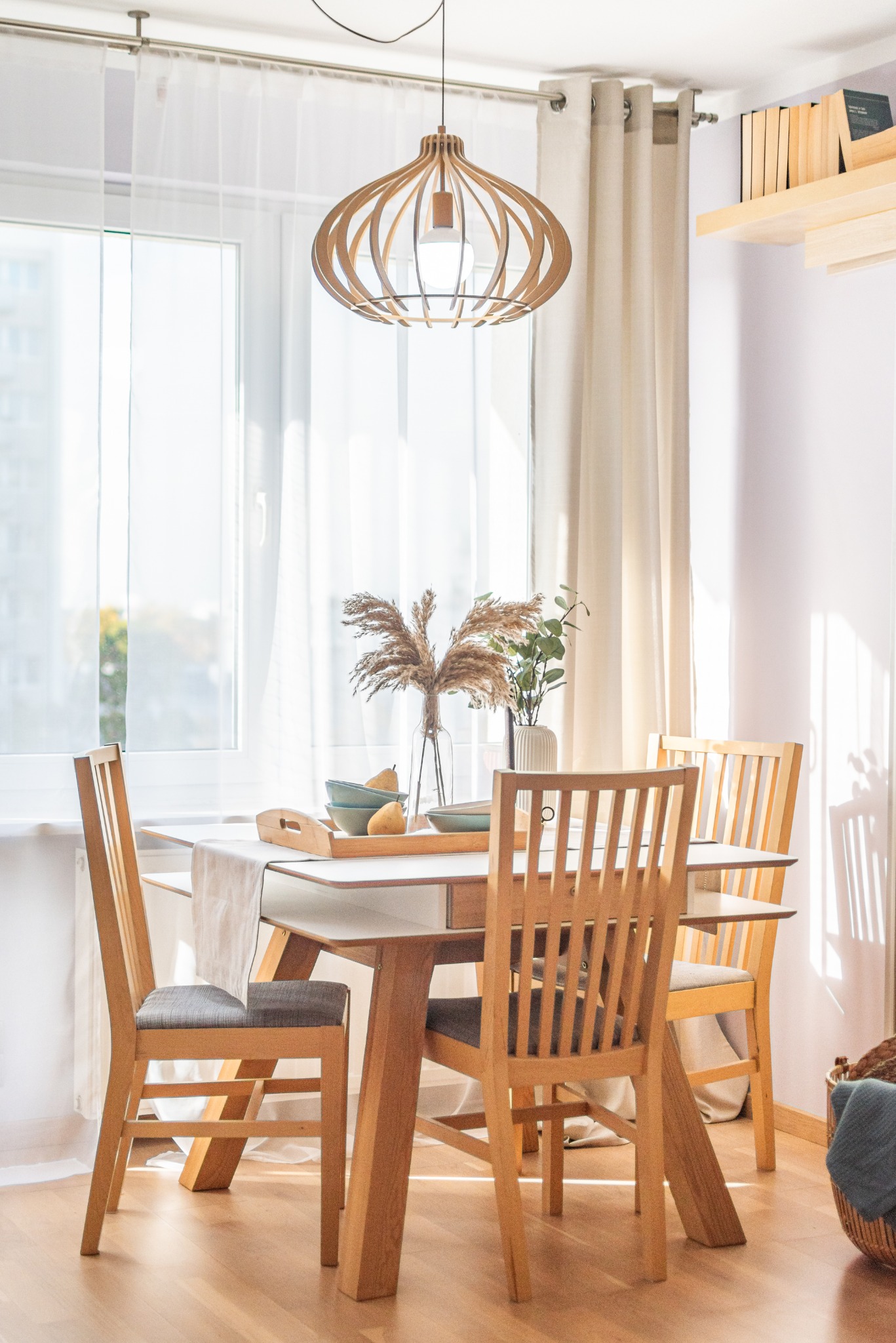 Jasne wnętrze jadalni z drewnianym stołem, krzesłami z szarymi poduszkami, dekoracyjną lampą wiszącą i bukietami suszonych traw w wazonach, oświetlone promieniami słońca wpadającymi przez okno...