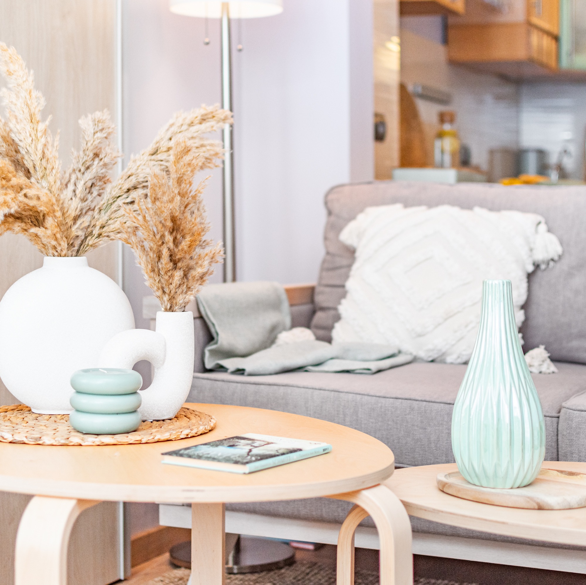 Jasne wnętrze: okrągłe stoliki kawowe z dekoracjami (wazony z suszem, świeczki, książka), szara sofa z poduszką i pledem, oświetlenie podłogowe.