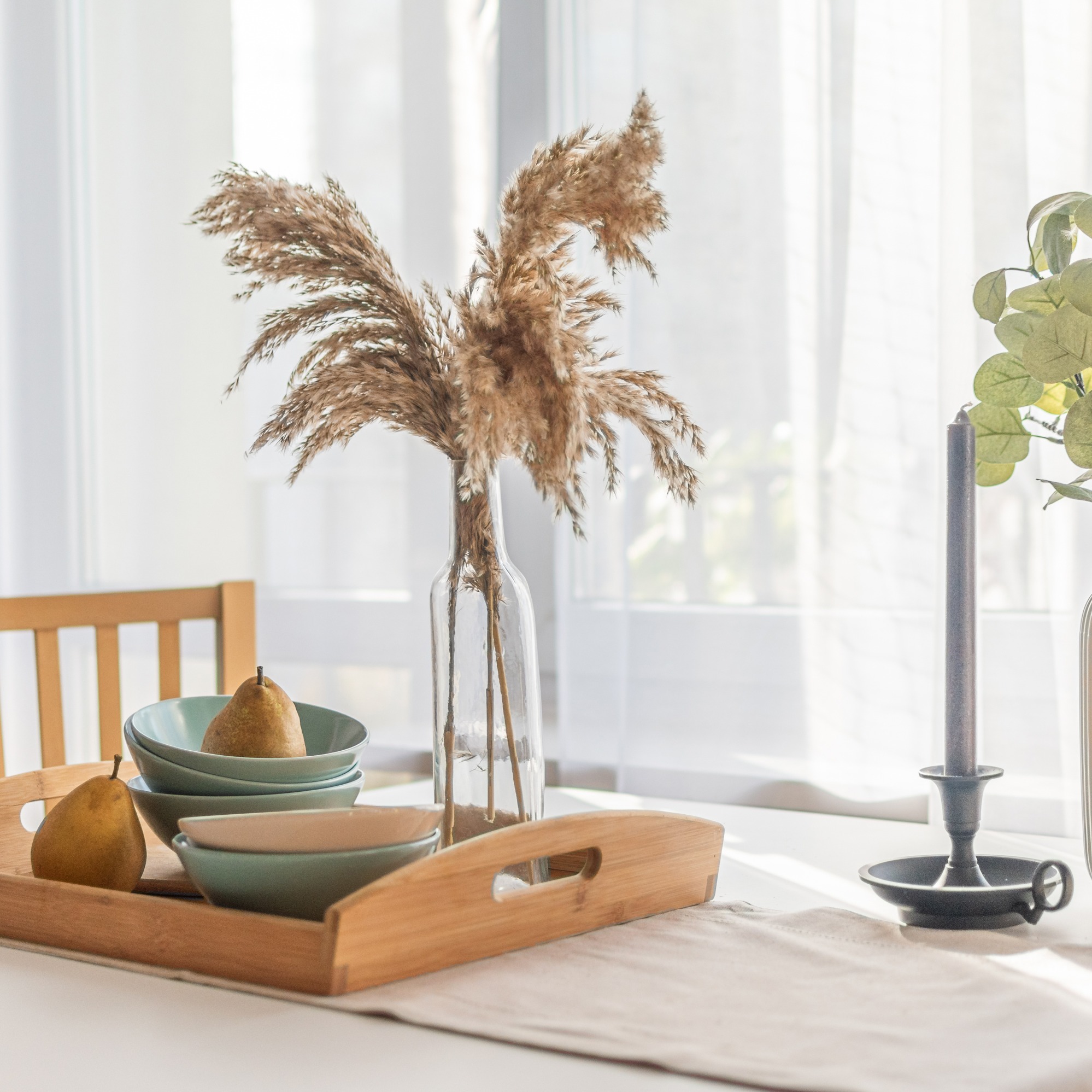 Stół z dekoracyjnym bukietem z traw w szklanym wazonie, drewnianą tacą z miseczkami i gruszkami oraz świecznikiem z szarą świecą, w tle okno z białymi zasłonami.