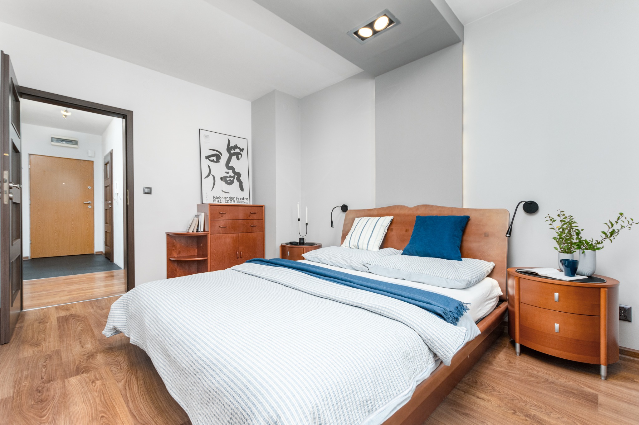 Jasna sypialnia z drewnianym łóżkiem z niebieskimi akcentami, drewnianymi szafkami nocnymi i komodą, plakat na ścianie, widok na otwarte drzwi do przedpokoju.