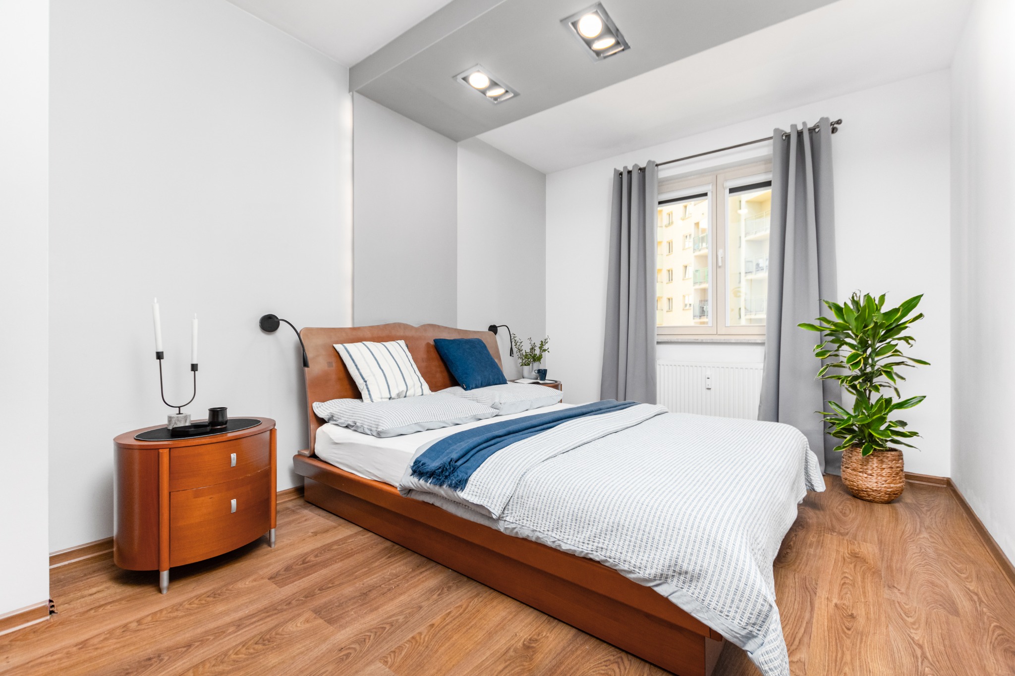 Jasna sypialnia z drewnianym łóżkiem, stolikiem nocnym z lampką i rośliną doniczkową przy oknie z szarymi zasłonami.