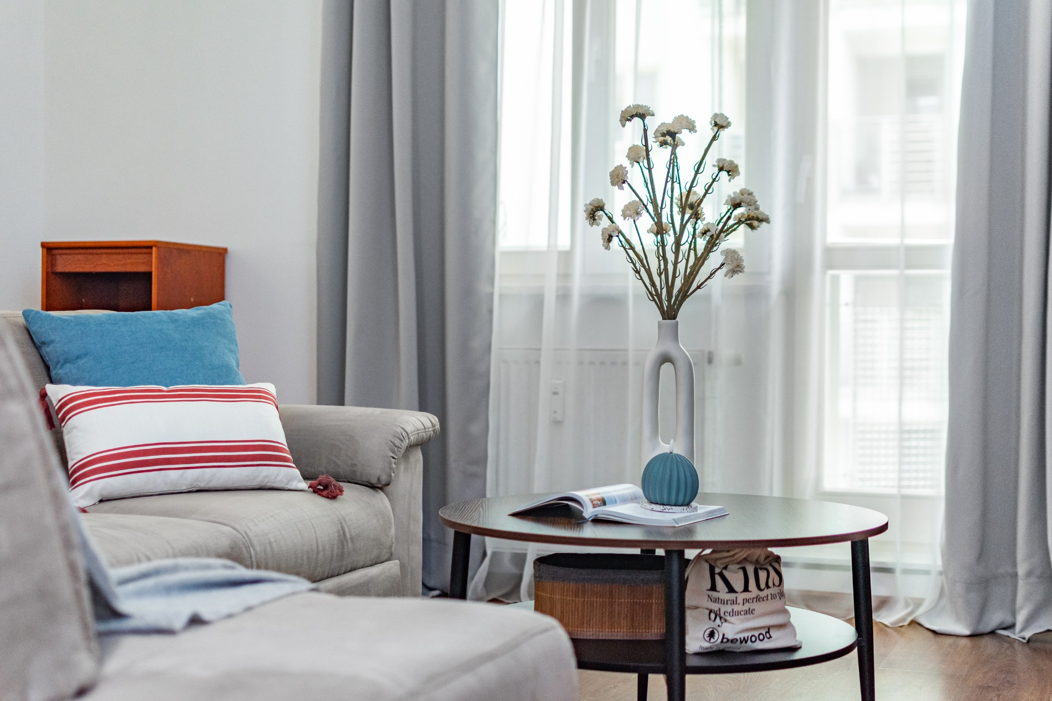 Jasny salon z beżową sofą z poduszkami w kolorze niebieskim i białym z czerwonymi paskami, okrągłym stolikiem kawowym z wazonem z białymi kwiatami, otwartą książką i dekoracyjną niebieską kulą...