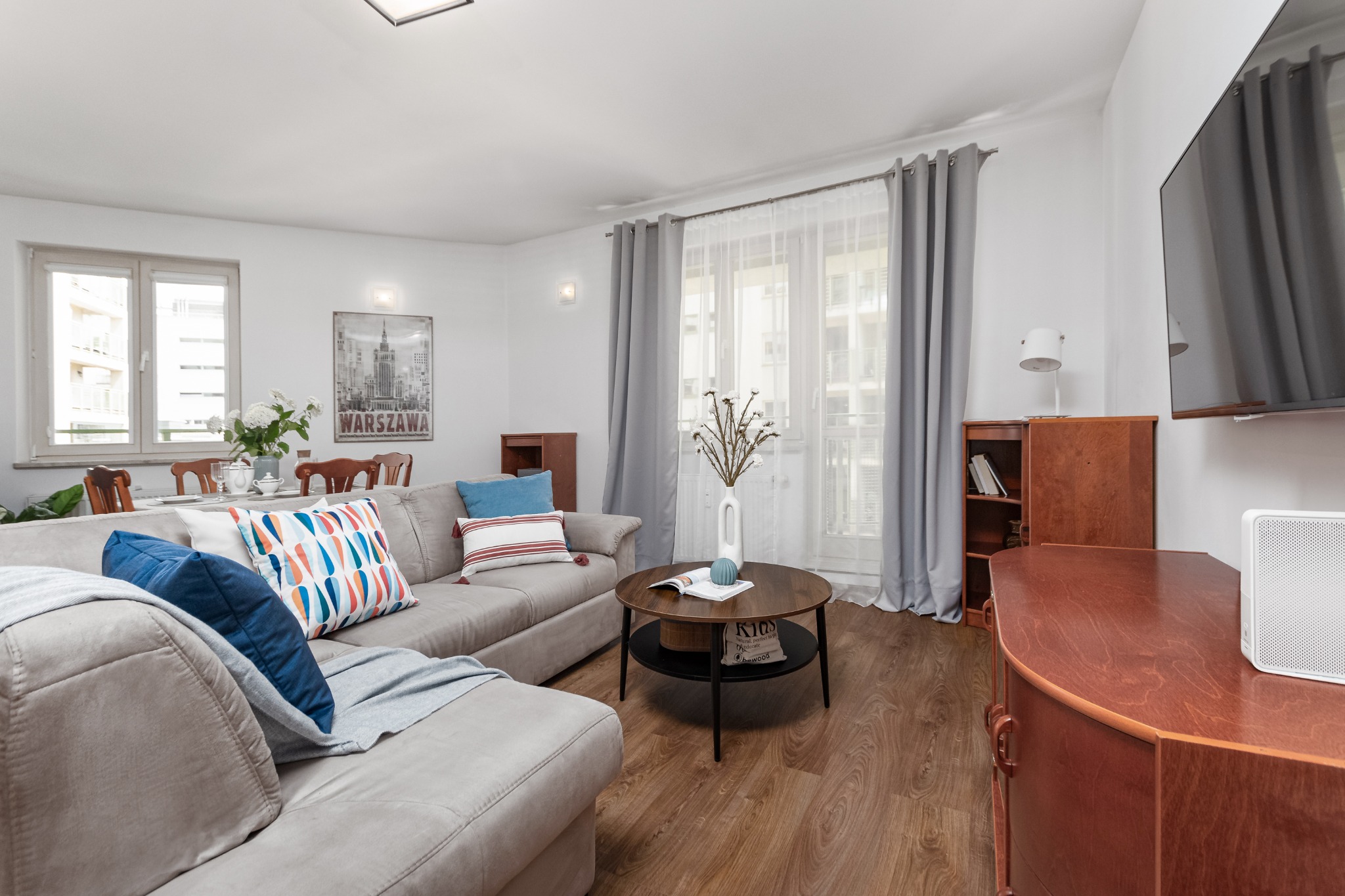 Jasne, przestronne wnętrze salonu z szarą sofą narożną, drewnianym stolikiem kawowym i plakat z motywem Warszawy na ścianie.