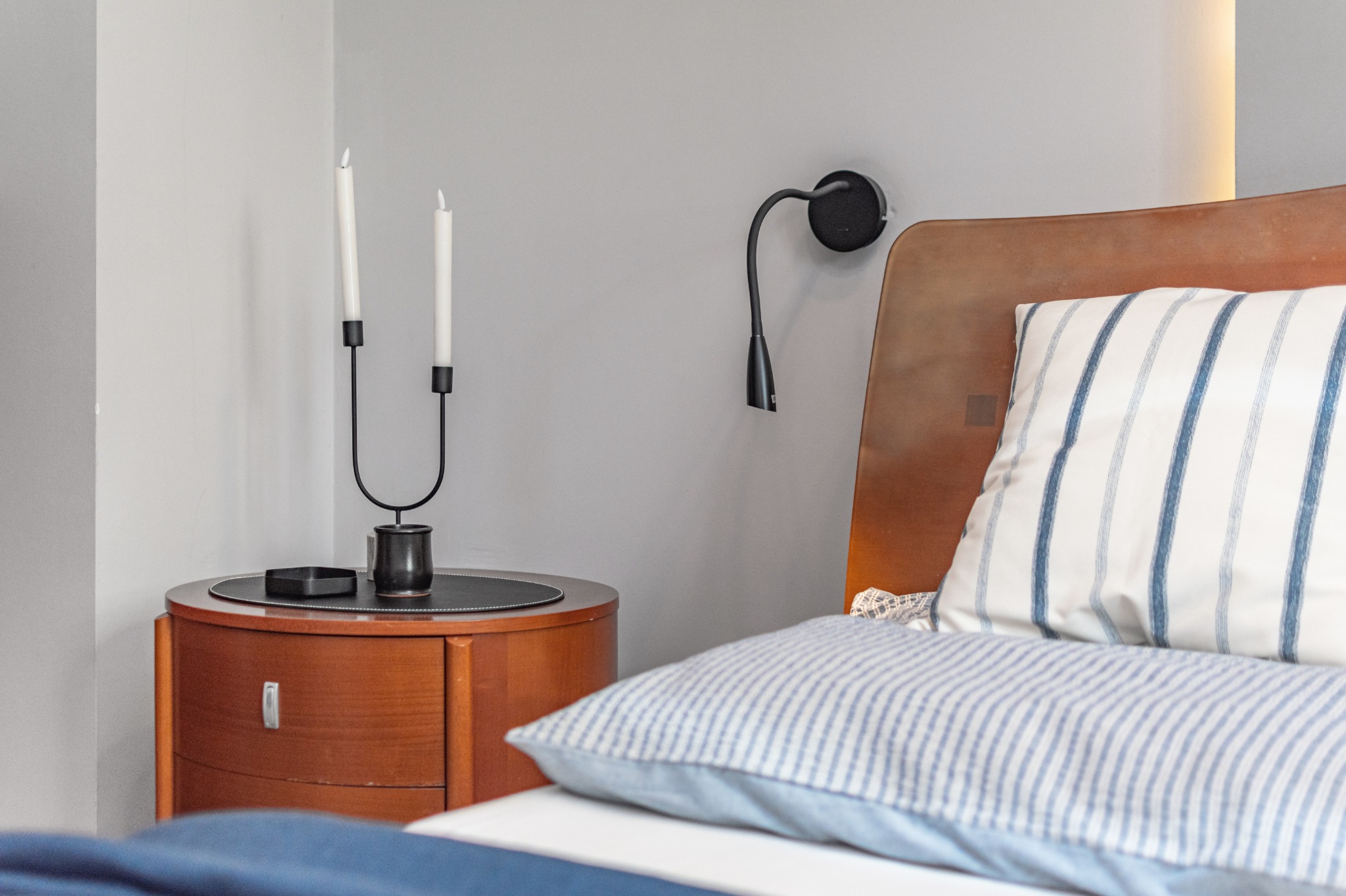 Sypialnia z drewnianym łóżkiem i stolikiem nocnym w stylu retro z czarnym świecznikiem na dwie świece i lampką ścienną.