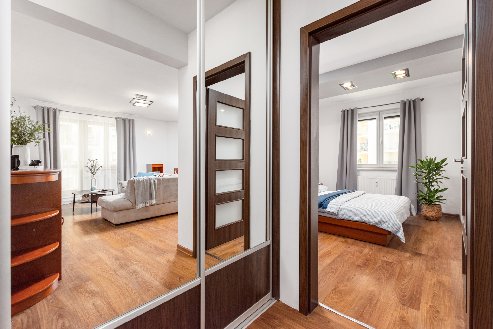 Widok na salon i sypialnię poprzez otwarte drzwi i lustrzane drzwi szafy, ukazujący aranżację wnętrz z drewnianą podłogą i elementami dekoracyjnymi.