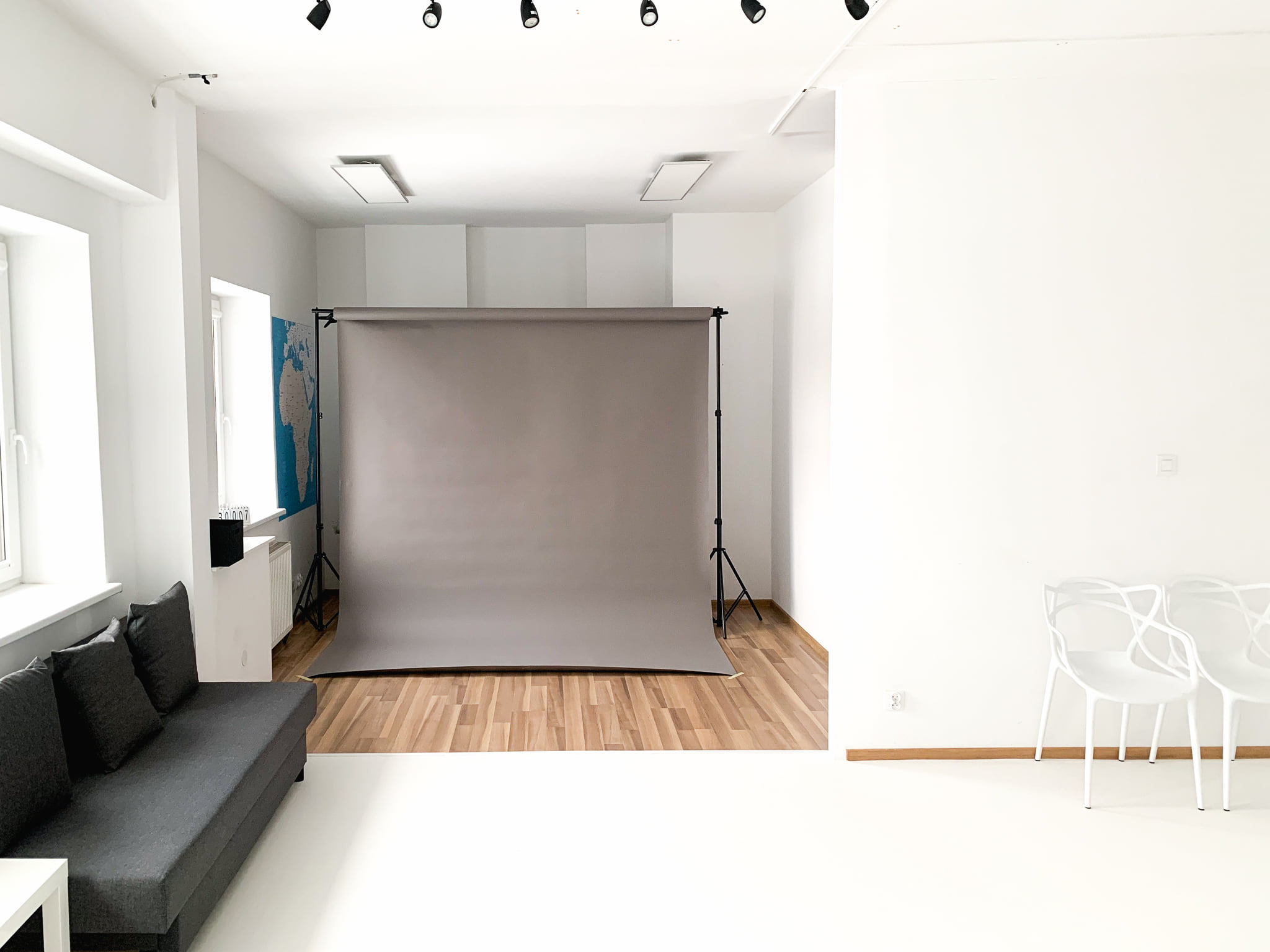 Jasne studio fotograficzne z szarym tłem, sofą, białymi krzesłami i oświetleniem sufitowym.