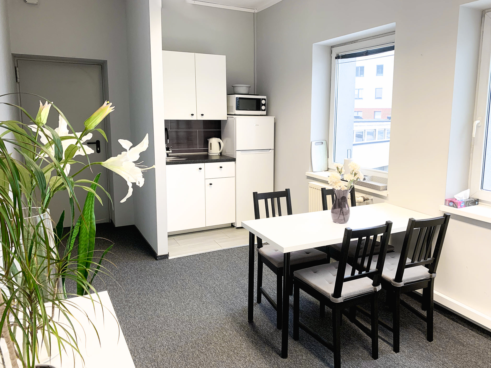 Jasne wnętrze pokoju z aneksem kuchennym, biały stół z czarnymi krzesłami, kwiaty w wazonie, minimalistyczny wystrój.