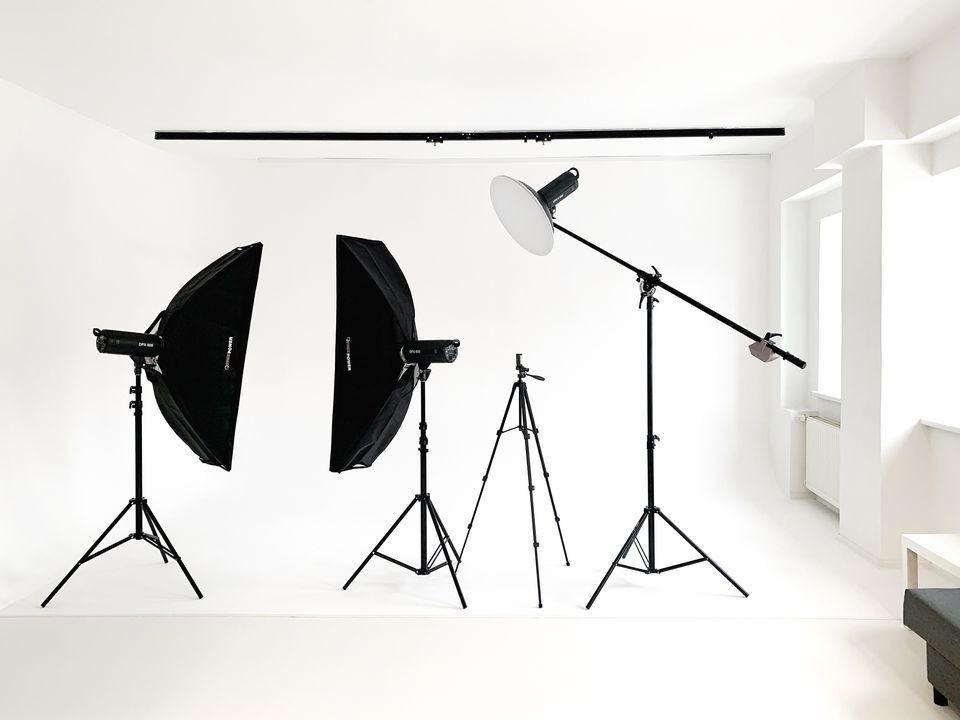 Puste studio fotograficzne: dwa softboxy na statywach, reflektor na wysięgniku, statyw aparatu, białe ściany i podłoga.