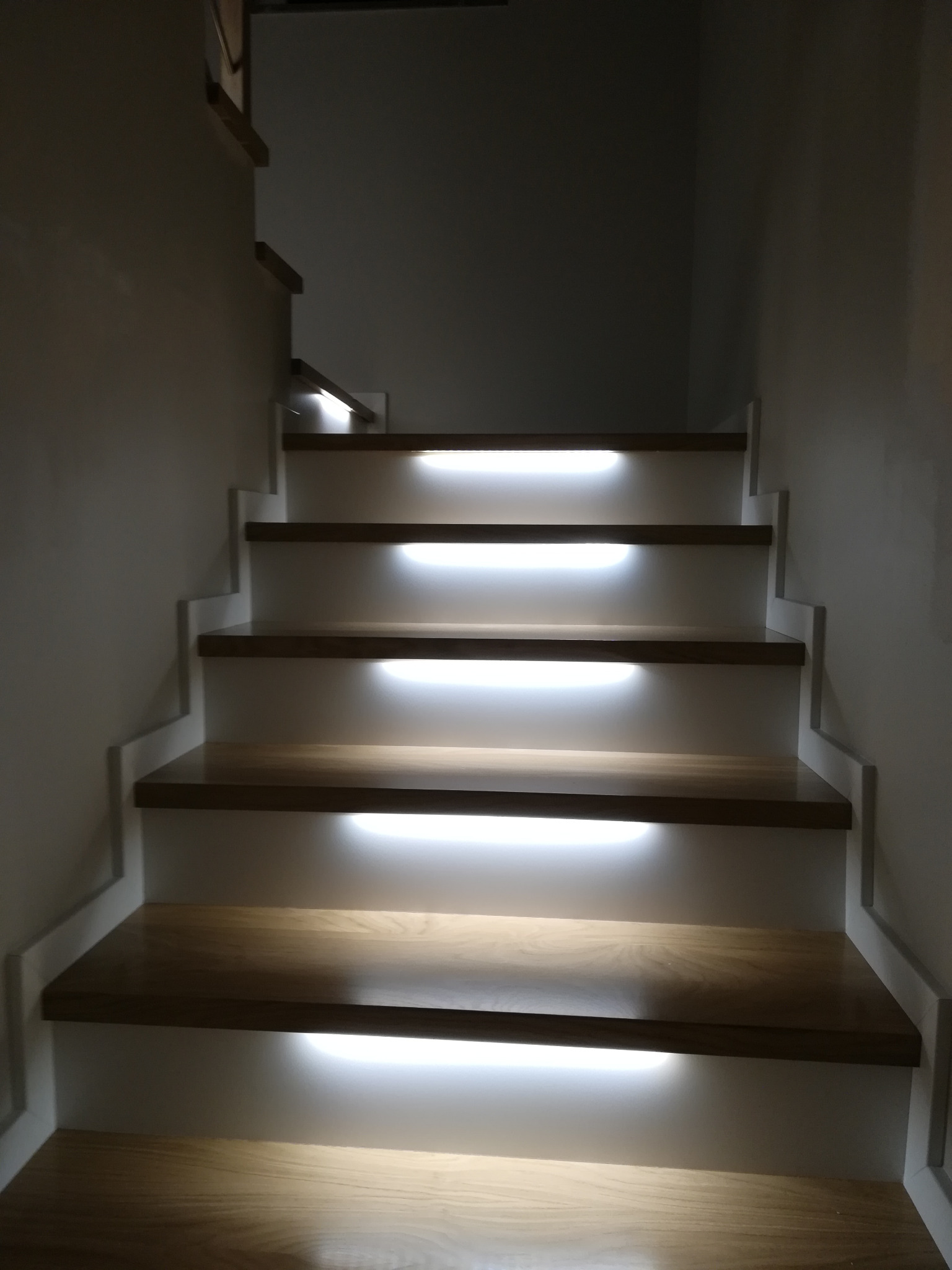 Oświetlone schody drewniane z białymi podstopnicami, zintegrowane oświetlenie LED pod każdym stopniem, widok z dołu w górę, minimalistyczny design.