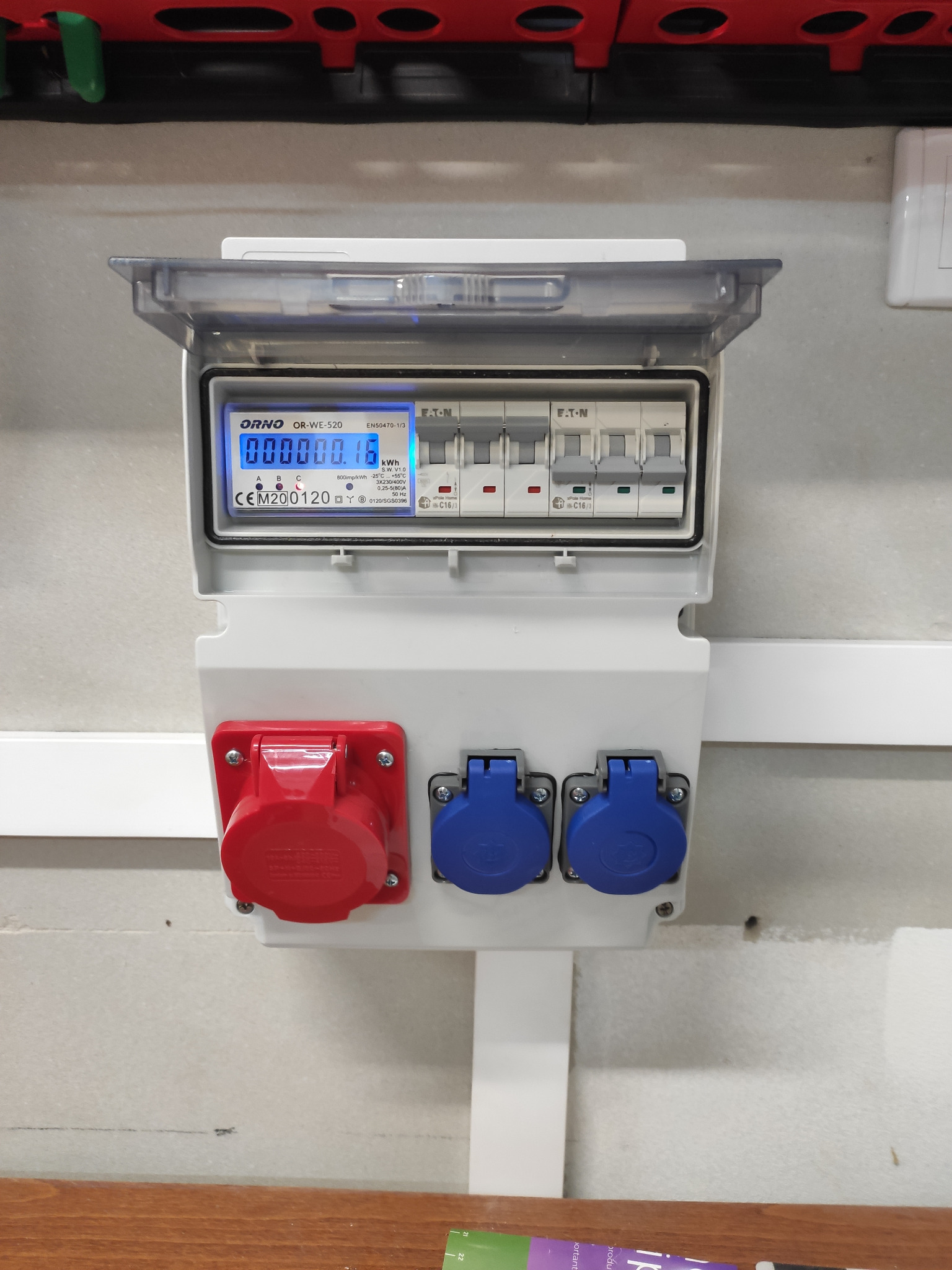 Szara skrzynka elektryczna z przezroczystą osłoną, zawierająca licznik energii Orno OR-WE-520 z wyświetlaczem LCD, zabezpieczenia nadprądowe Eaton oraz dwa niebieskie i jedno czerwone gniazdo siłowe.