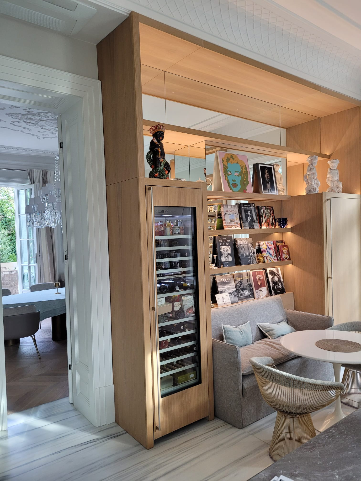 Drewniana zabudowa regału z półkami, wbudowaną chłodziarką na wino, szarą kanapą i okrągłym białym stolikiem z metalowymi krzesłami w jasnym wnętrzu.