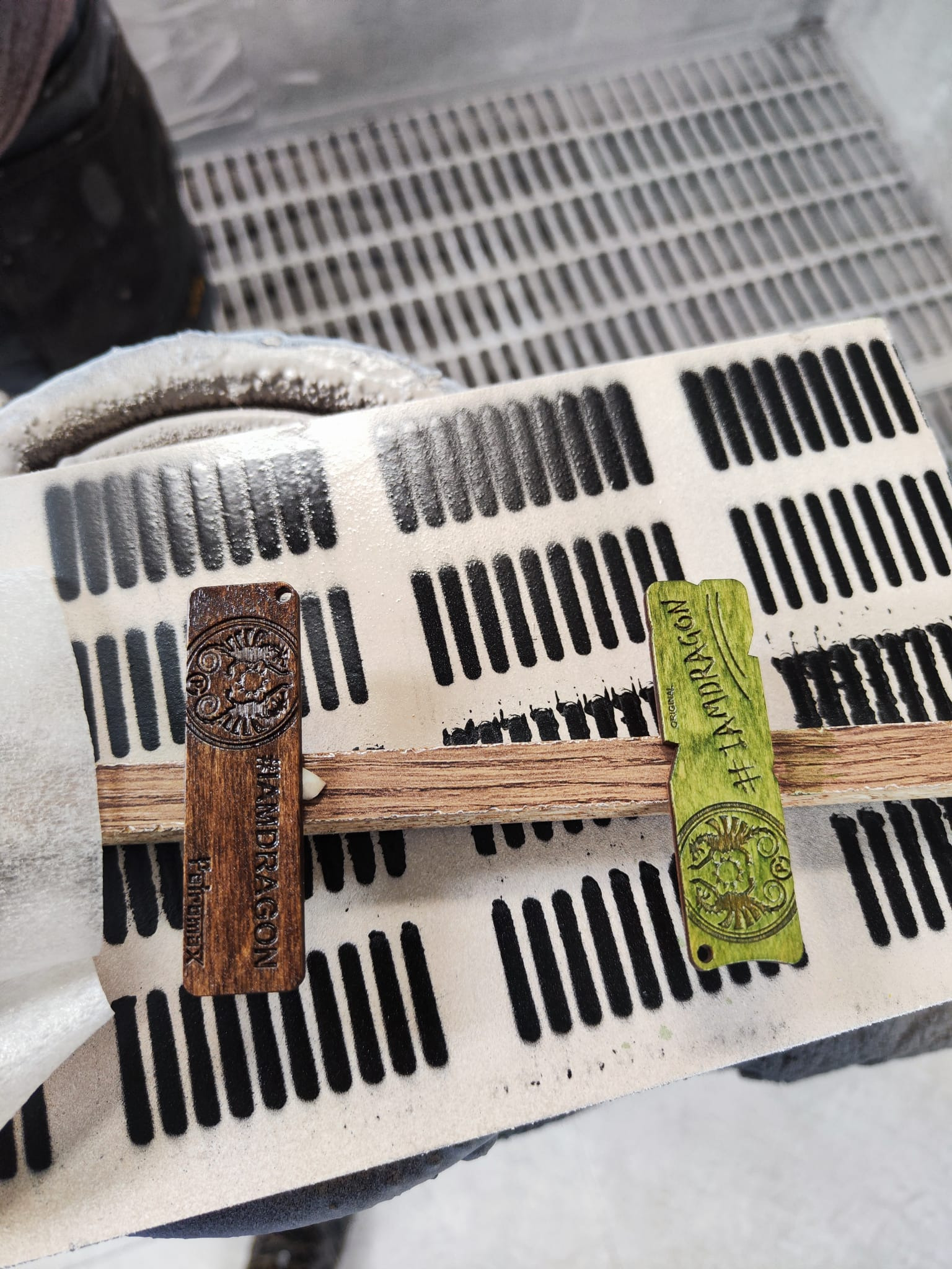 Dwa drewniane breloczki z wygrawerowanym wzorem i napisem #IAMDRAGON, jeden w kolorze naturalnego drewna, drugi pomalowany na zielono, leżące na lakierniczym stojaku.