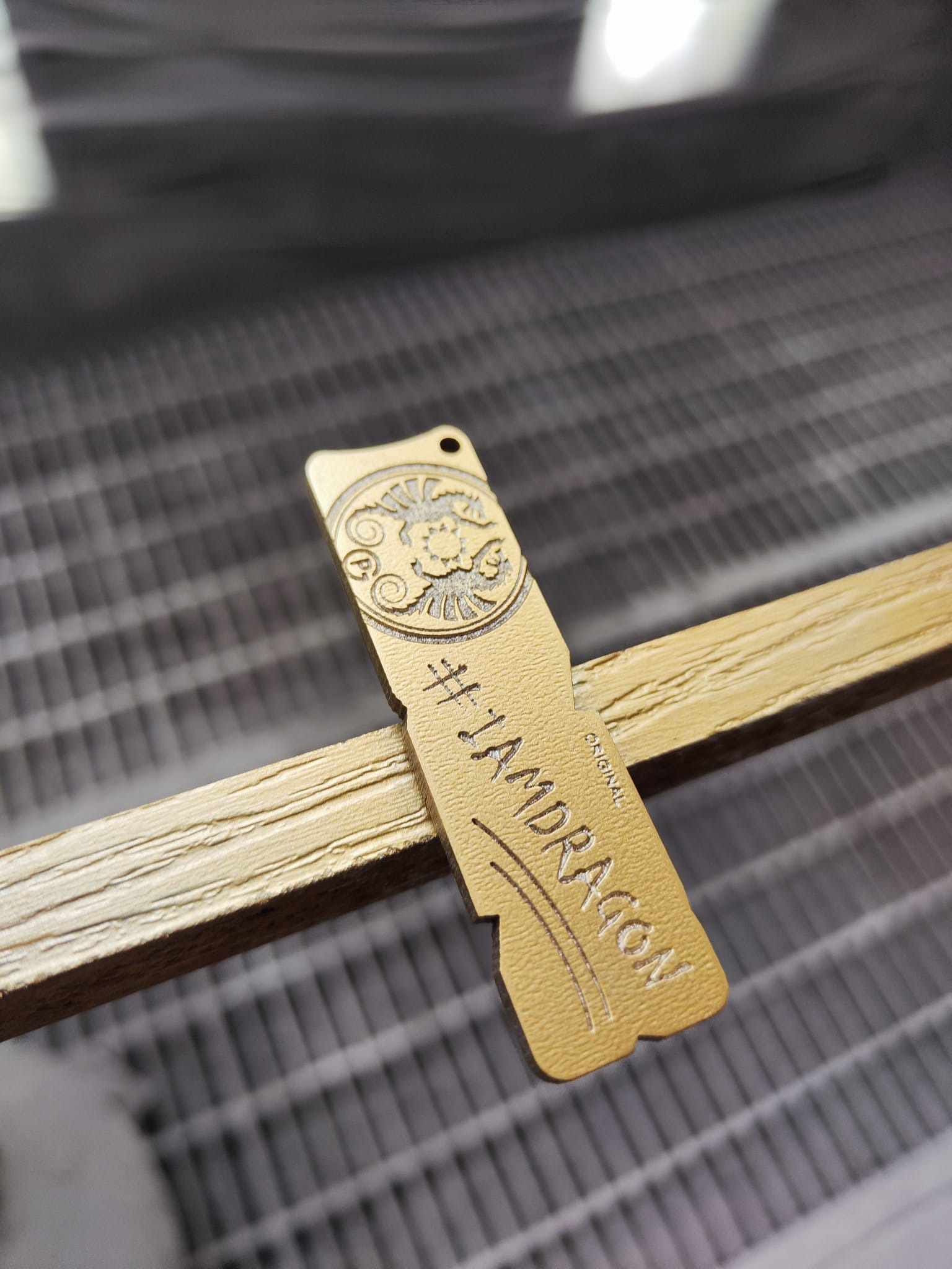 Drewniany brelok z wygrawerowanym wzorem kwiatowym i napisem '#IAMDRAGON ORIGINAL' leżący na drewnianej ramie z widocznym tłem kratki.