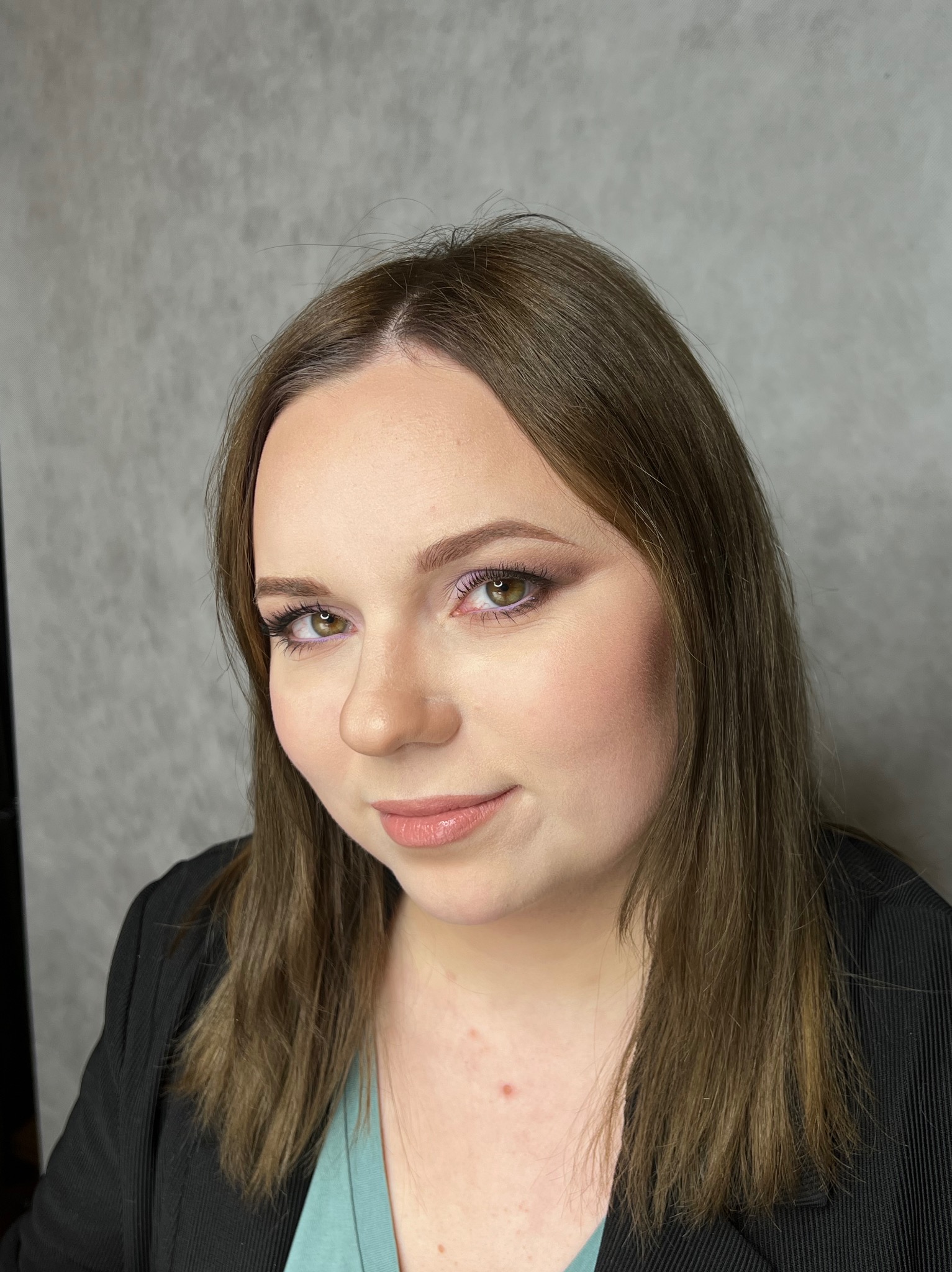 Portret kobiety z delikatnym, dziennym upiększeniem, z podkreślonymi oczami i ustami w odcieniach nude, na tle szarej ściany.