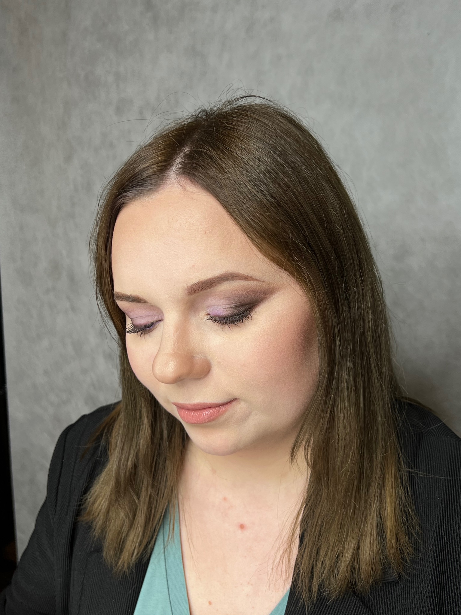 Portret kobiety z subtelnym, fioletowym cieniem na powiekach, delikatnym różem na policzkach i błyszczącymi ustami, skierowanej wzrokiem w dół.