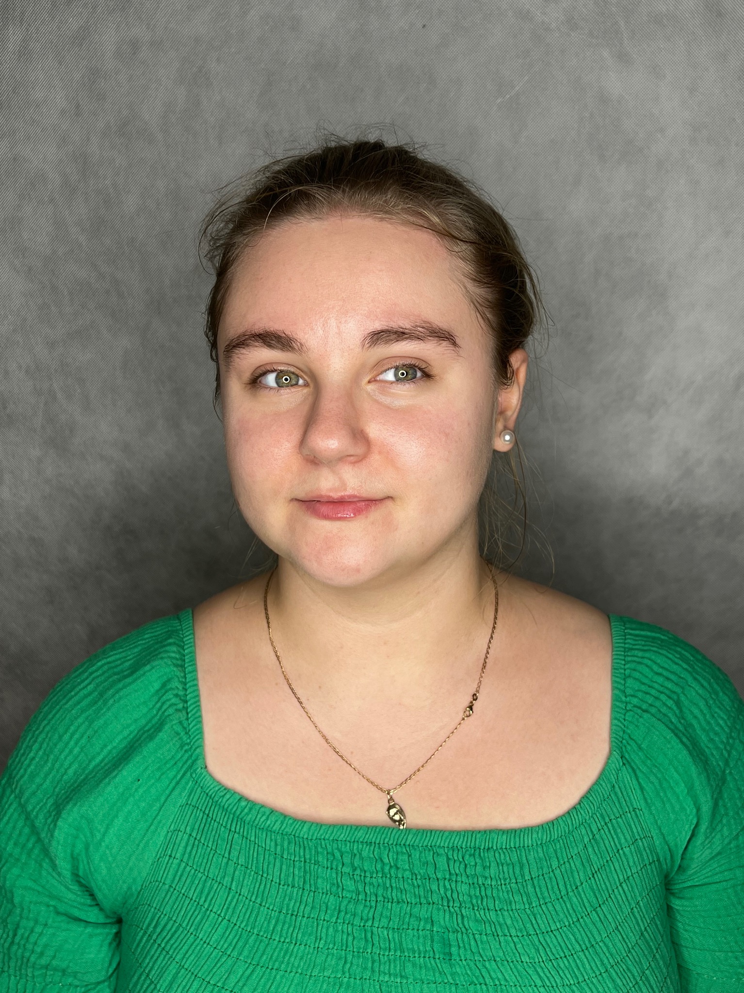 Portret kobiety z delikatnym makijażem brwi, ubranej w zieloną bluzkę, z widoczną złotą biżuterią na tle szarego tła.