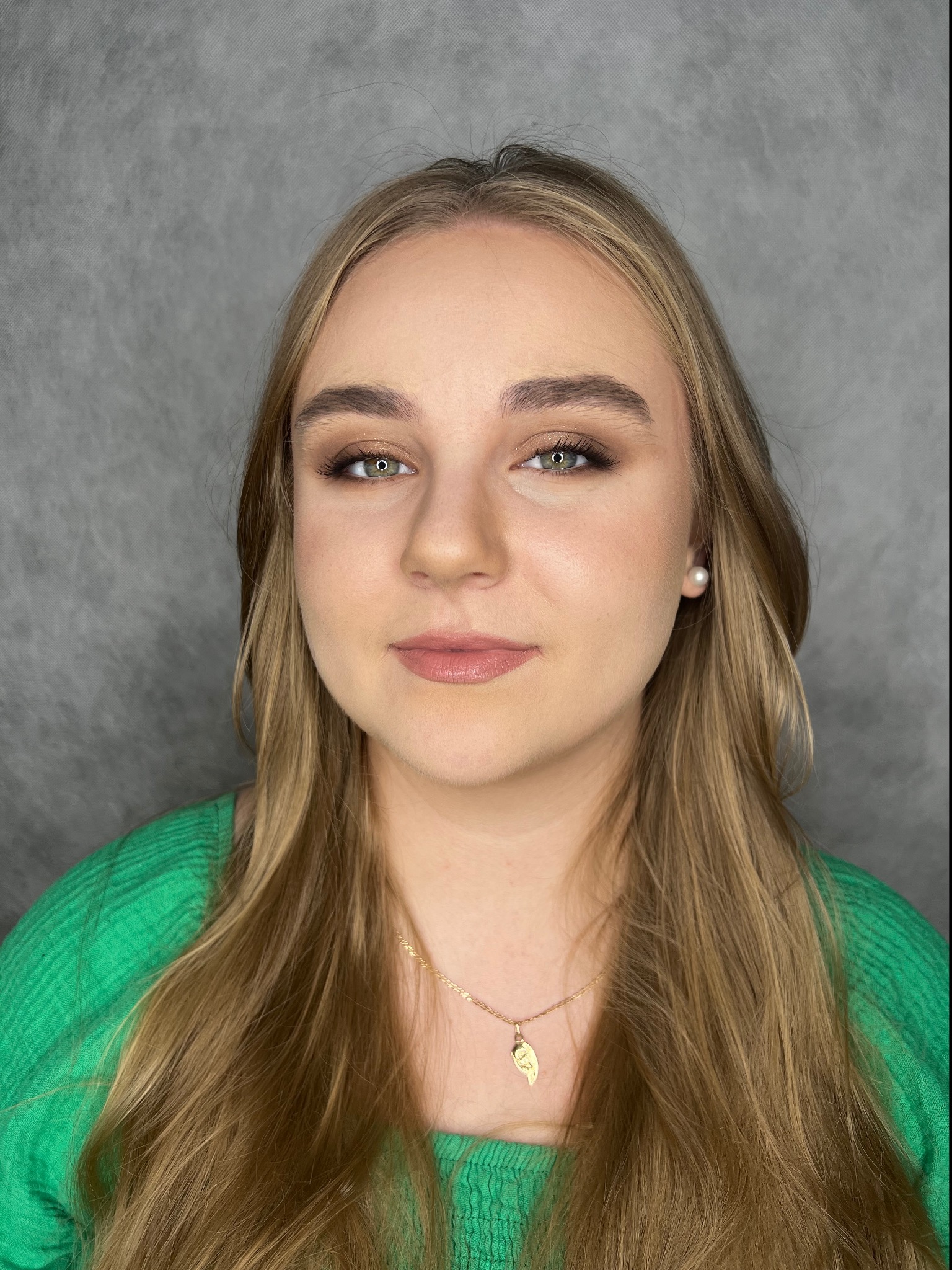 Portret młodej kobiety z delikatnym, brązowym cieniowaniem powiek, podkreślonymi brwiami i błyszczącymi ustami, uzupełniony perłowym kolczykiem i złotym naszyjnikiem na tle szarego tła.