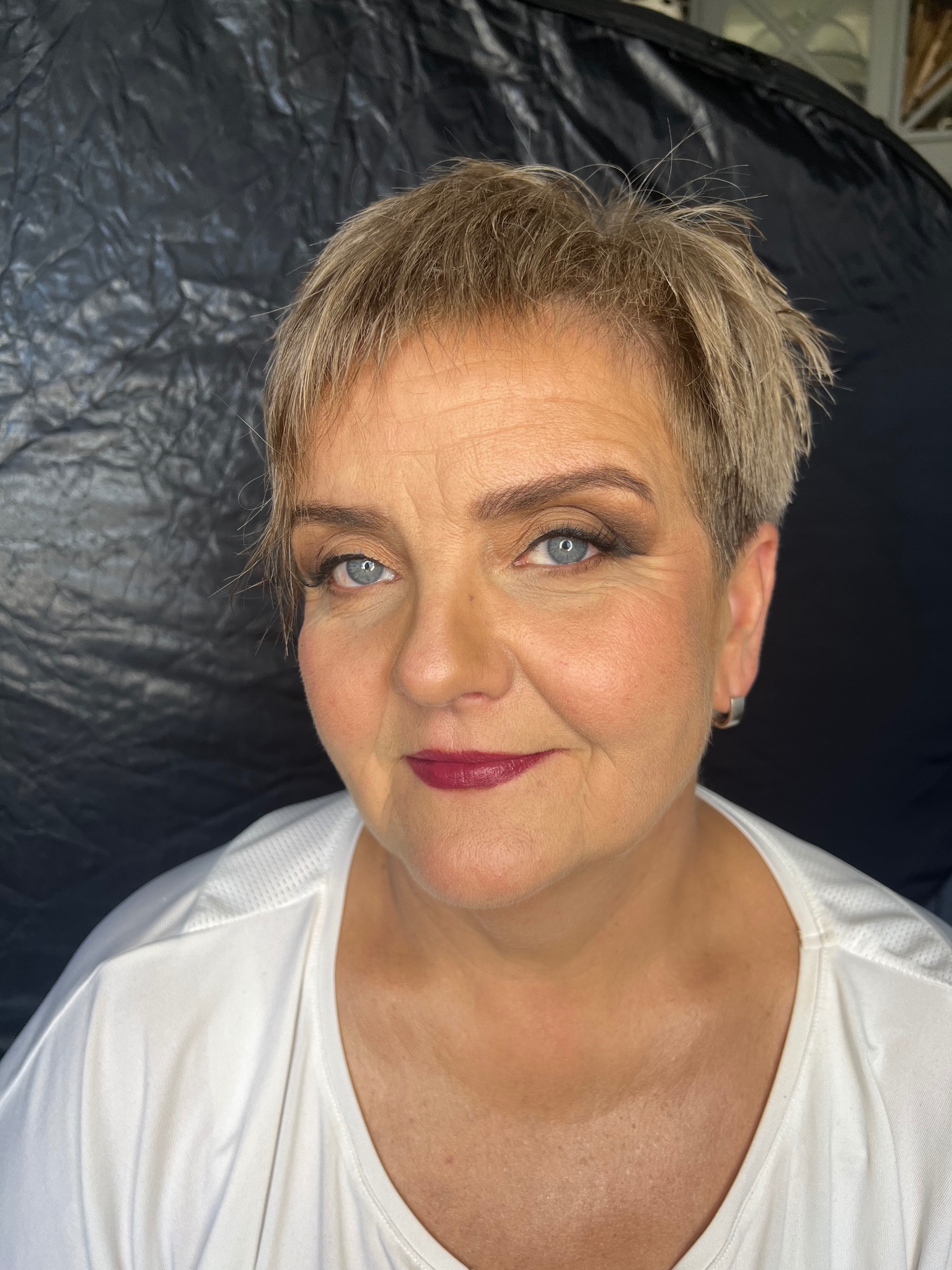 Portret kobiety z krótkimi blond włosami, w białej koszulce, z wykonanym makijażem oczu w odcieniach brązu i bordową pomadką na ustach, na czarnym tle.