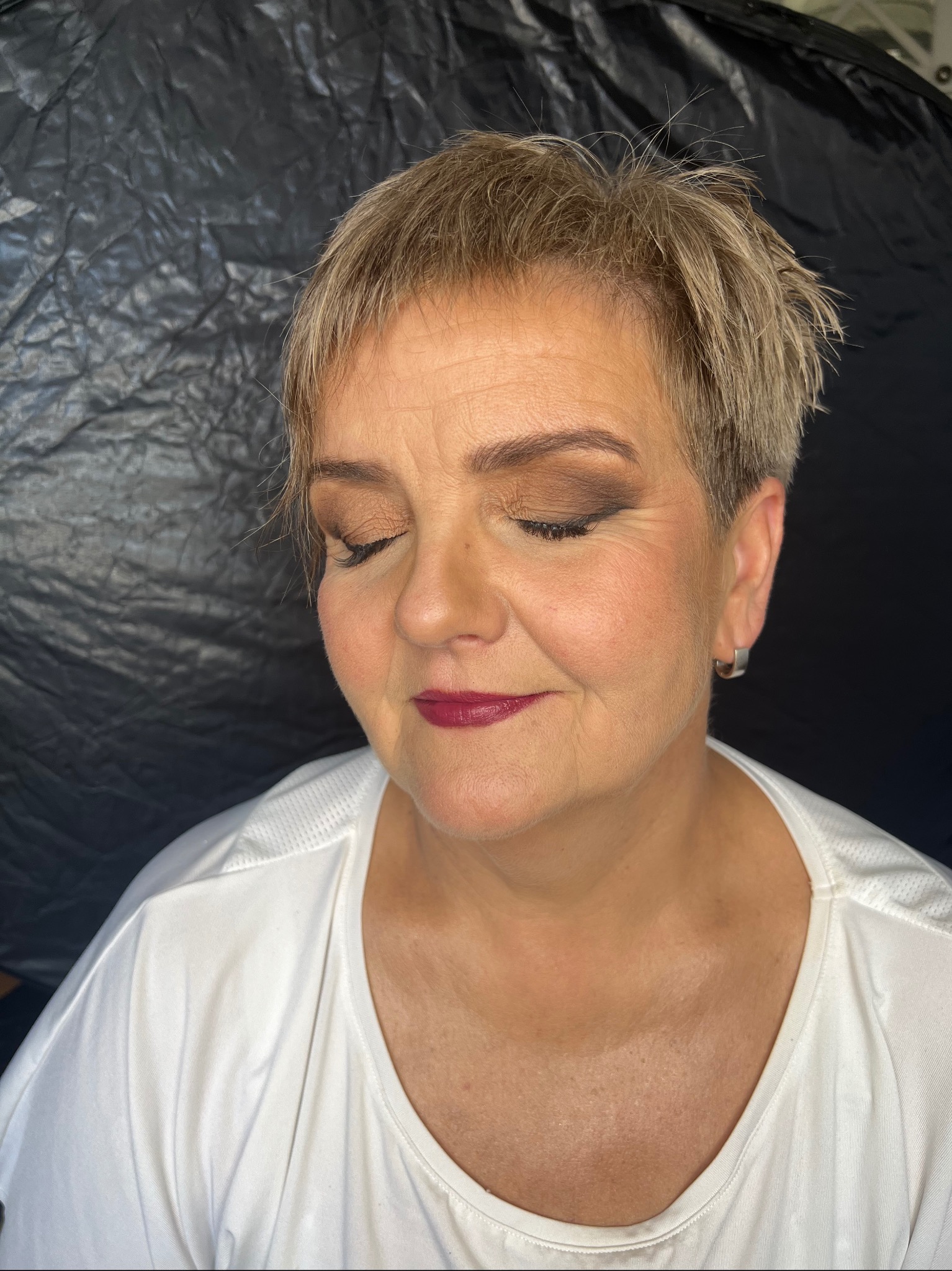 Portret kobiety z zamkniętymi oczami, z delikatnym brązowym cieniem na powiekach i bordową pomadką na ustach, na tle czarnego, pogniecionego materiału.