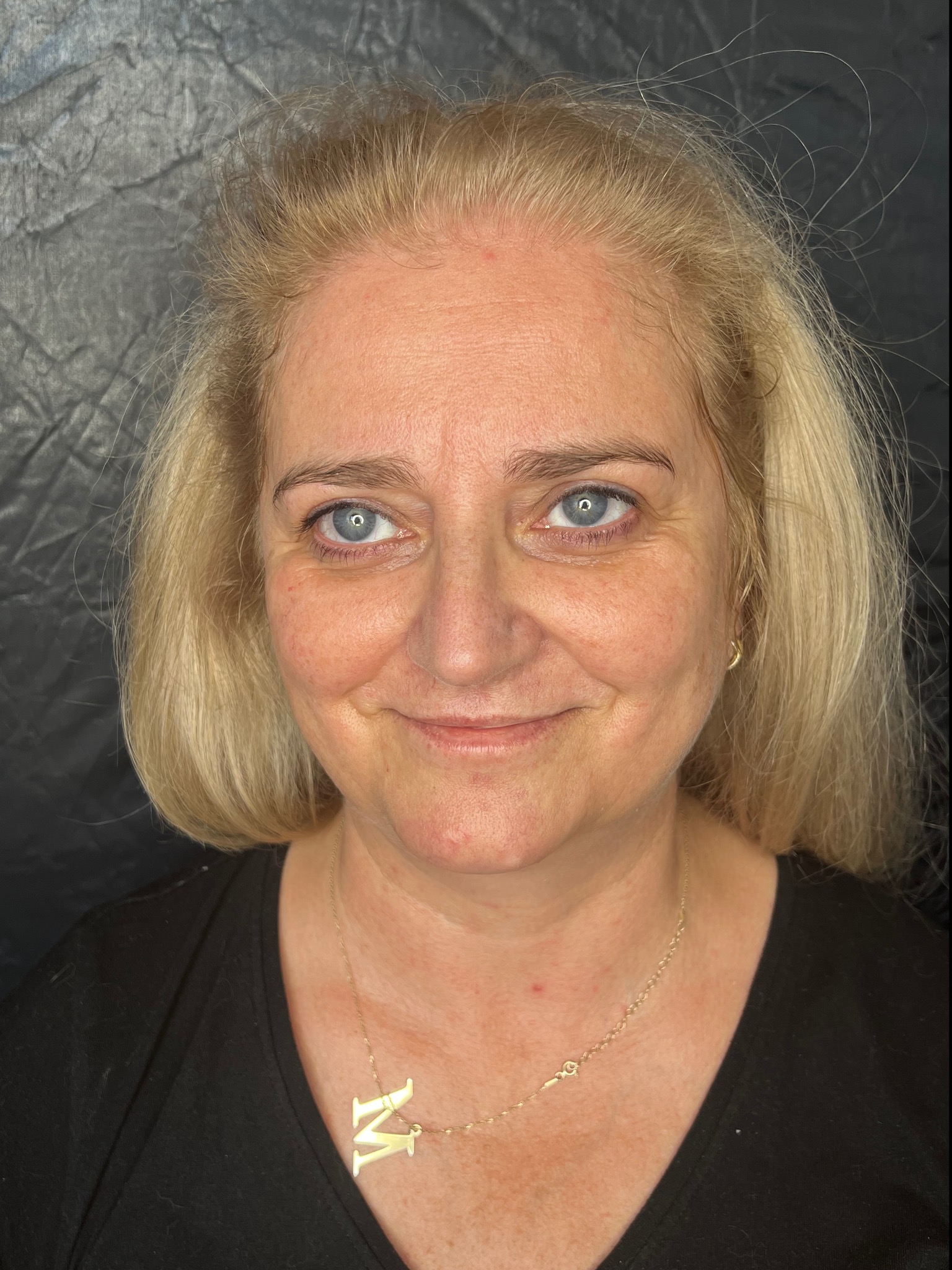 Portret kobiety o jasnych włosach i niebieskich oczach, uśmiechającej się do kamery, z widoczną złotą biżuterią na szyi, na tle ciemnego materiału.