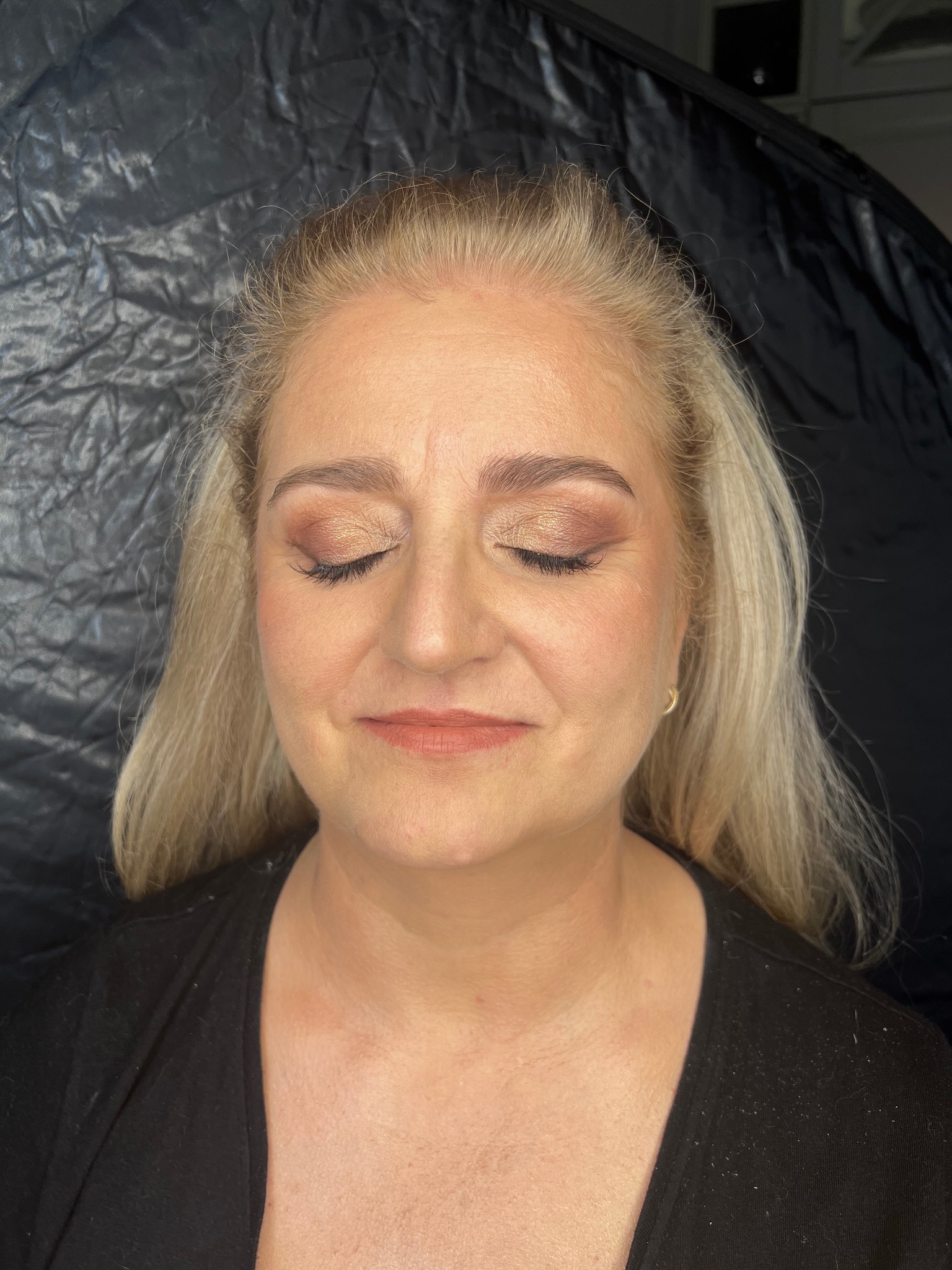 Portret kobiety z zamkniętymi oczami, prezentujący delikatny, złoty odcień cieni do powiek i subtelnie podkreślone brwi, na tle czarnej, lekko pogniecionej tkaniny.