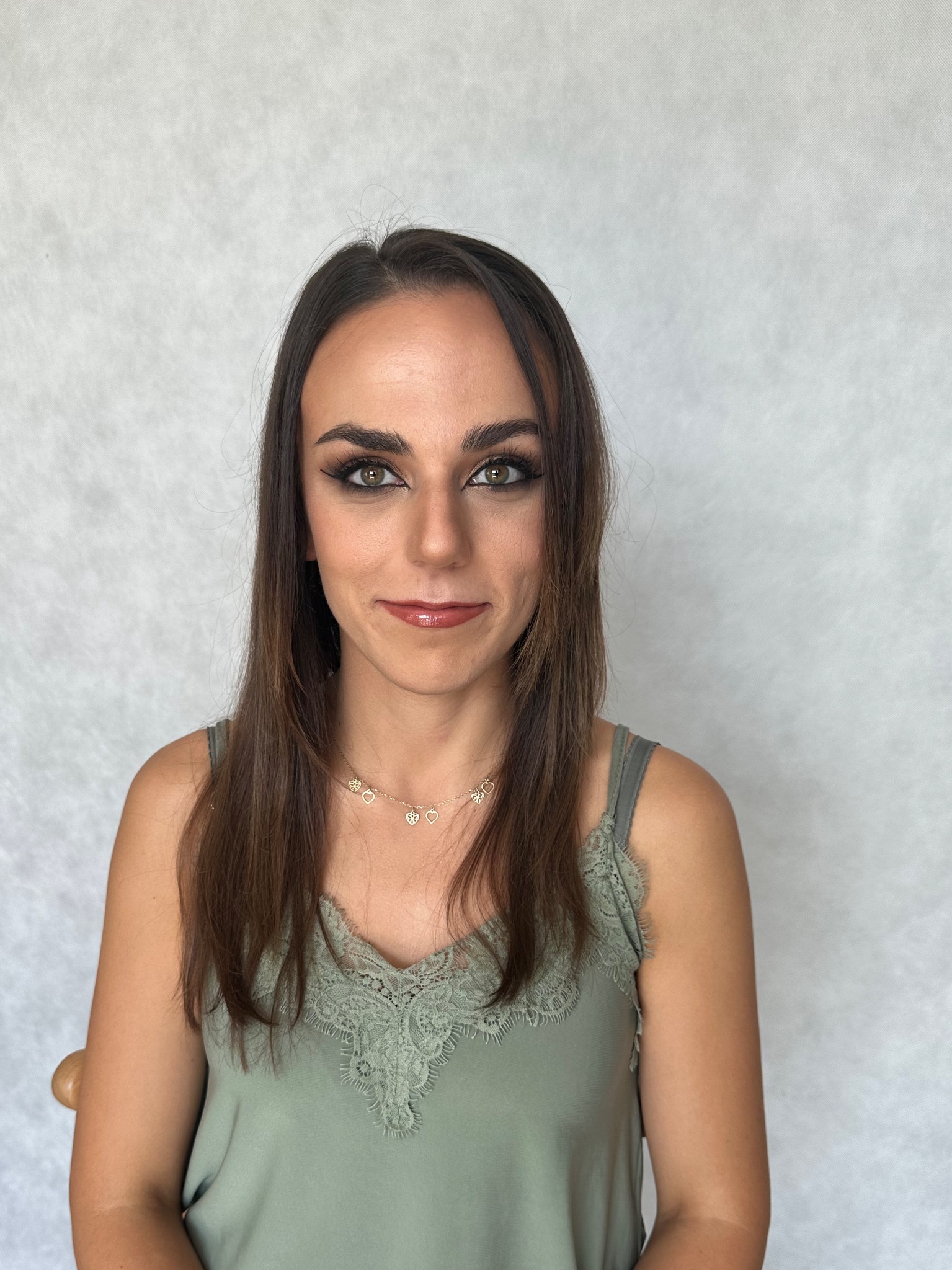 Portret kobiety z delikatnym, wieczorowym lookiem, podkreślone oczy cieniem i eyelinerem, błyszczące usta, włosy rozpuszczone, ubrana w zieloną bluzkę na ramiączkach z koronką i delikatny naszyjnik.